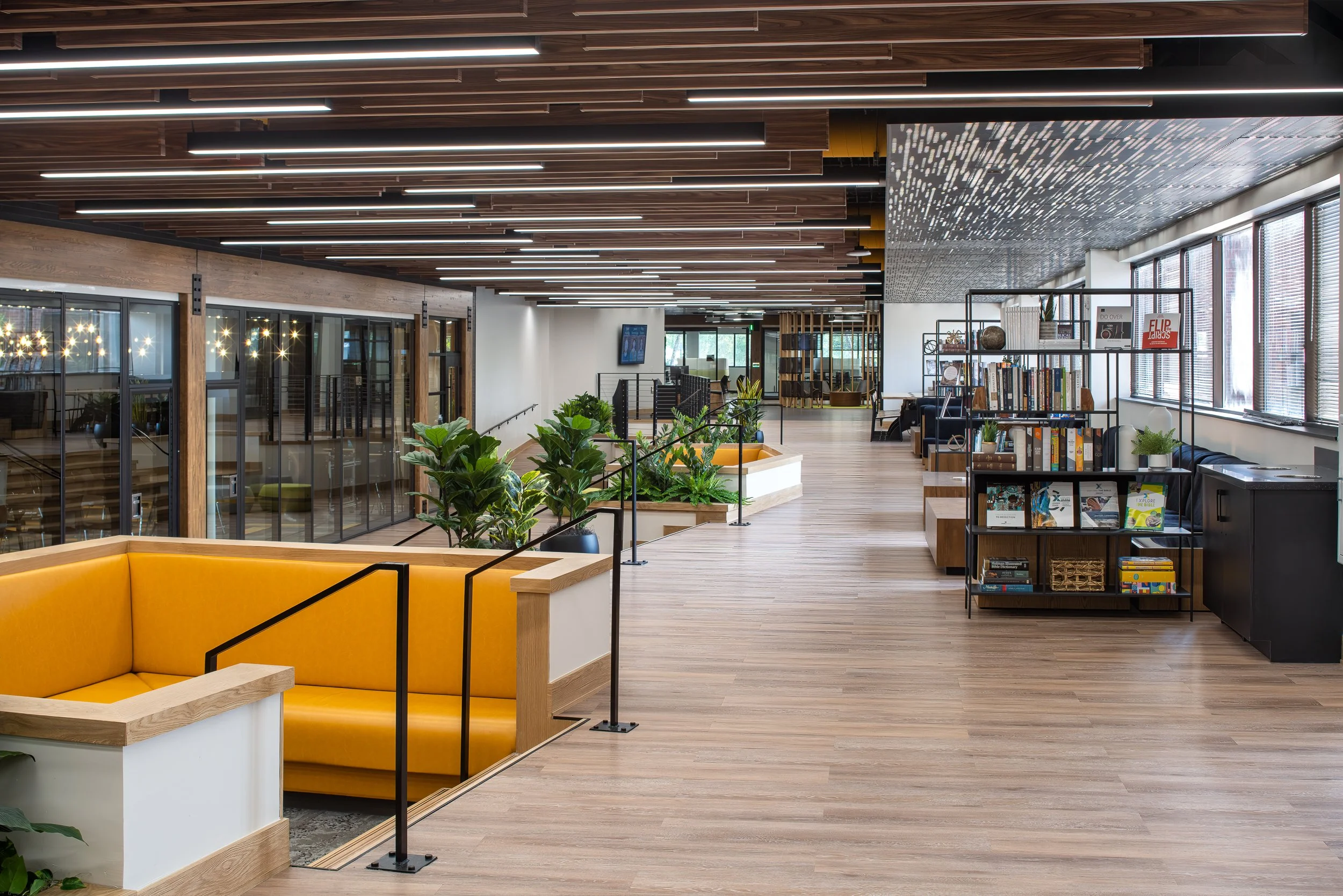 Modern office interior with wooden ceiling panels, large windows, indoor plants, and a bookshelf with various books, with a seating area featuring a yellow booth in the foreground.