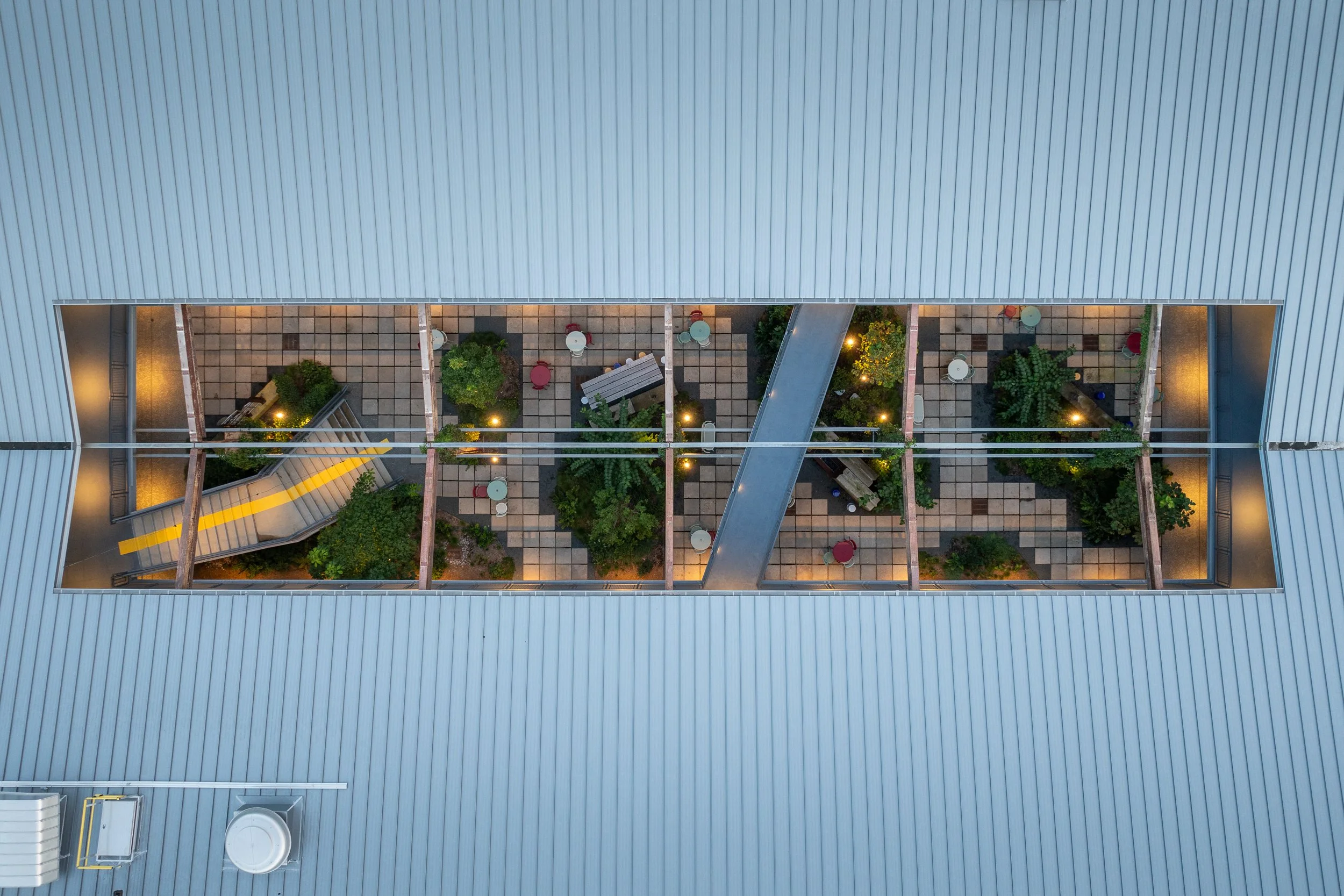 Aerial view looking down through the building's roof opening to the central courtyard, revealing the pattern of pavers, plantings, bistro tables, and the yellow-striped stair connecting the two levels.