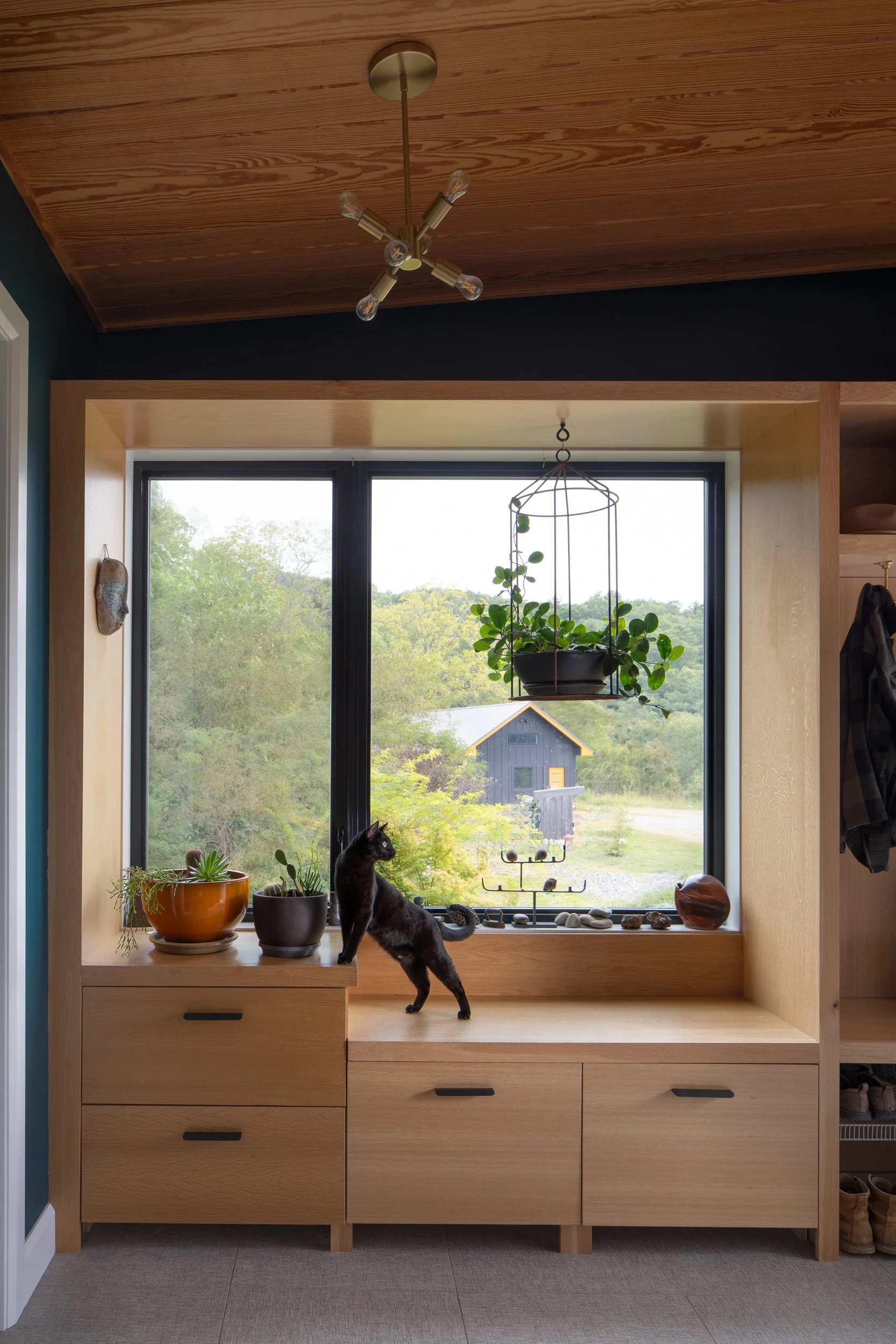 Black cat on a wooden built-in bench with potted plants near a window.