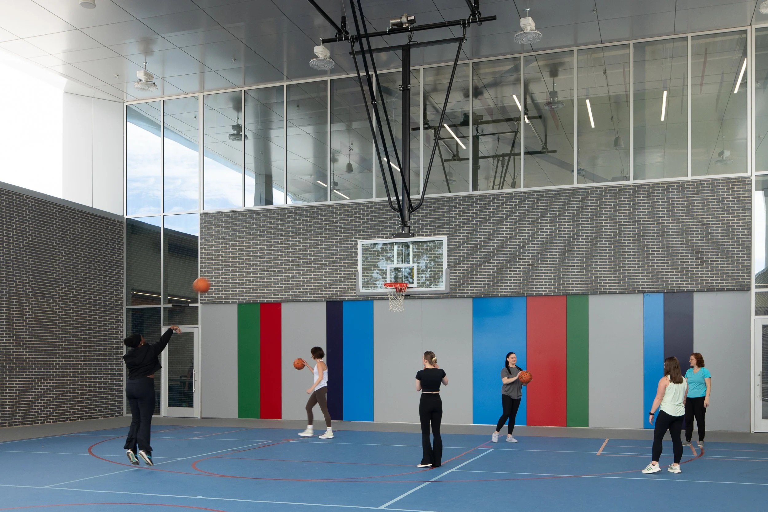 Basketball players practice on a covered blue court with tall glazing and vertical color panels behind the hoop.
