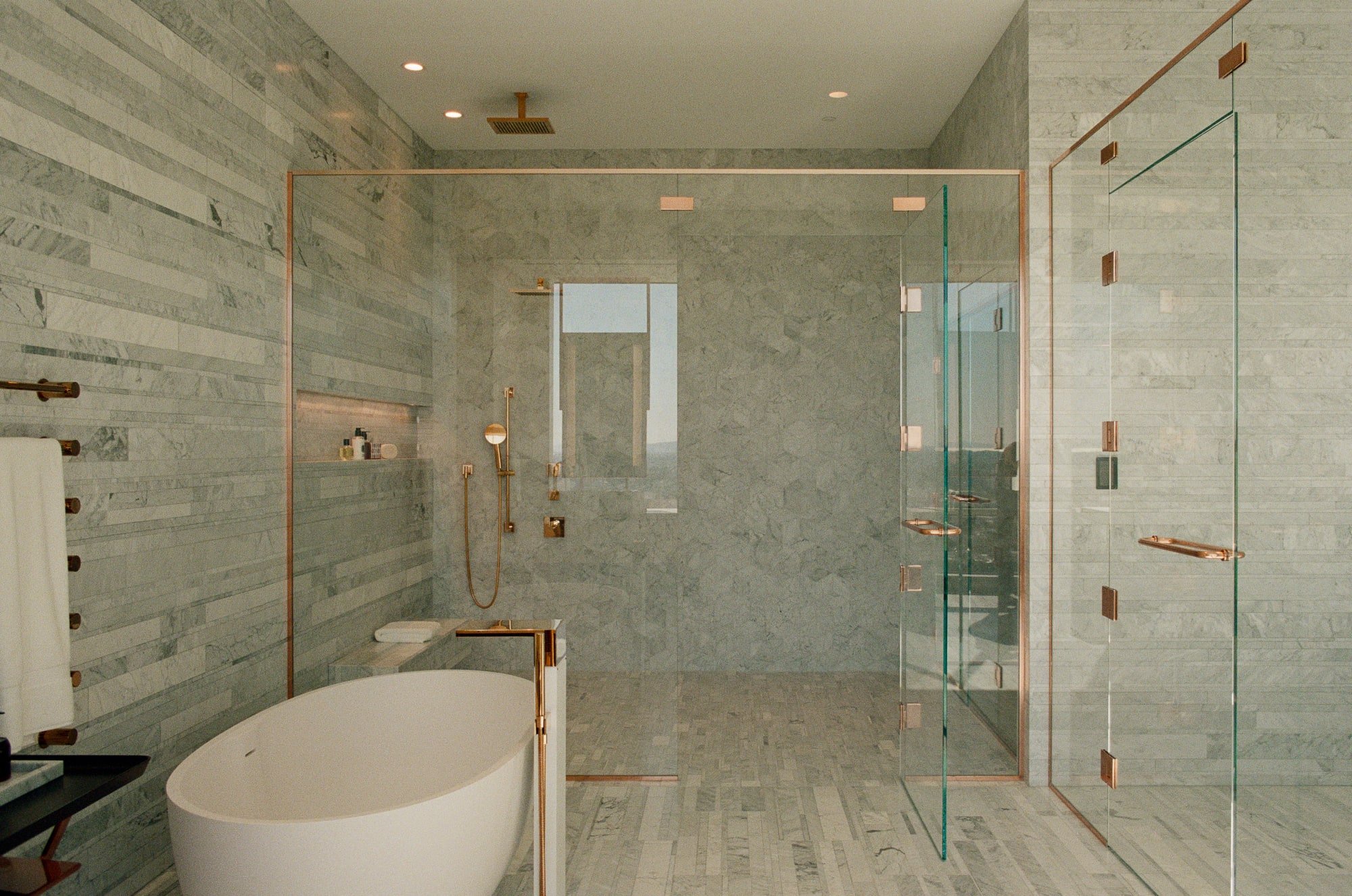Primary bathroom with freestanding tub, glass shower enclosure, marble tile, and copper-toned plumbing fixtures.