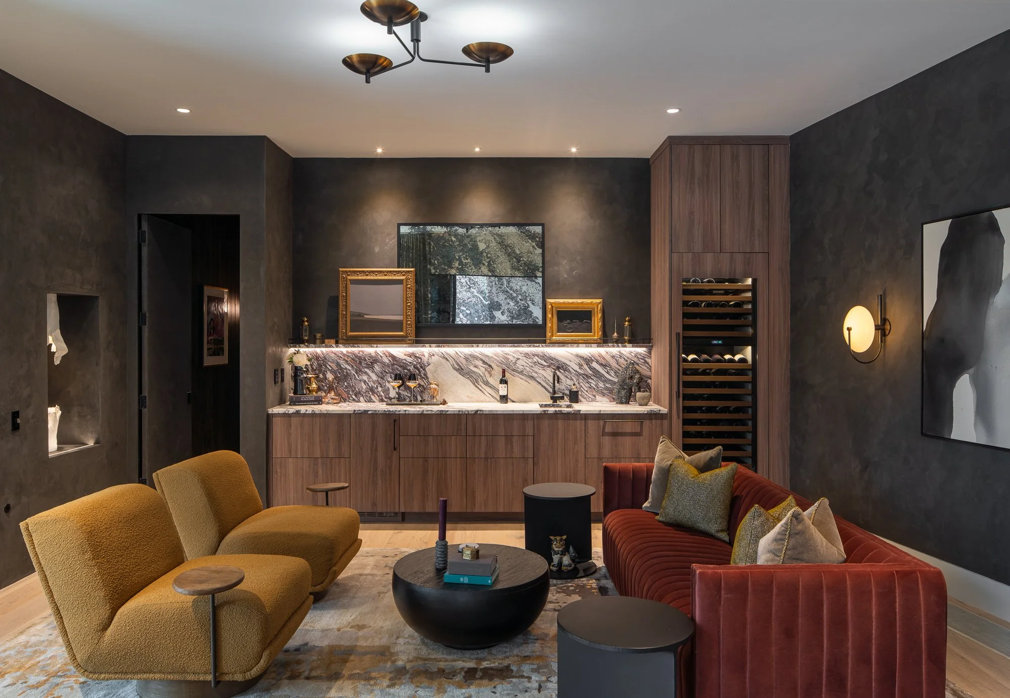 Living room featuring a bar area with a marble countertop, some framed art, a wine fridge, and modern seating including yellow and red velvet sofas with pillows, a black coffee table, and decorative lamps on textured dark walls.