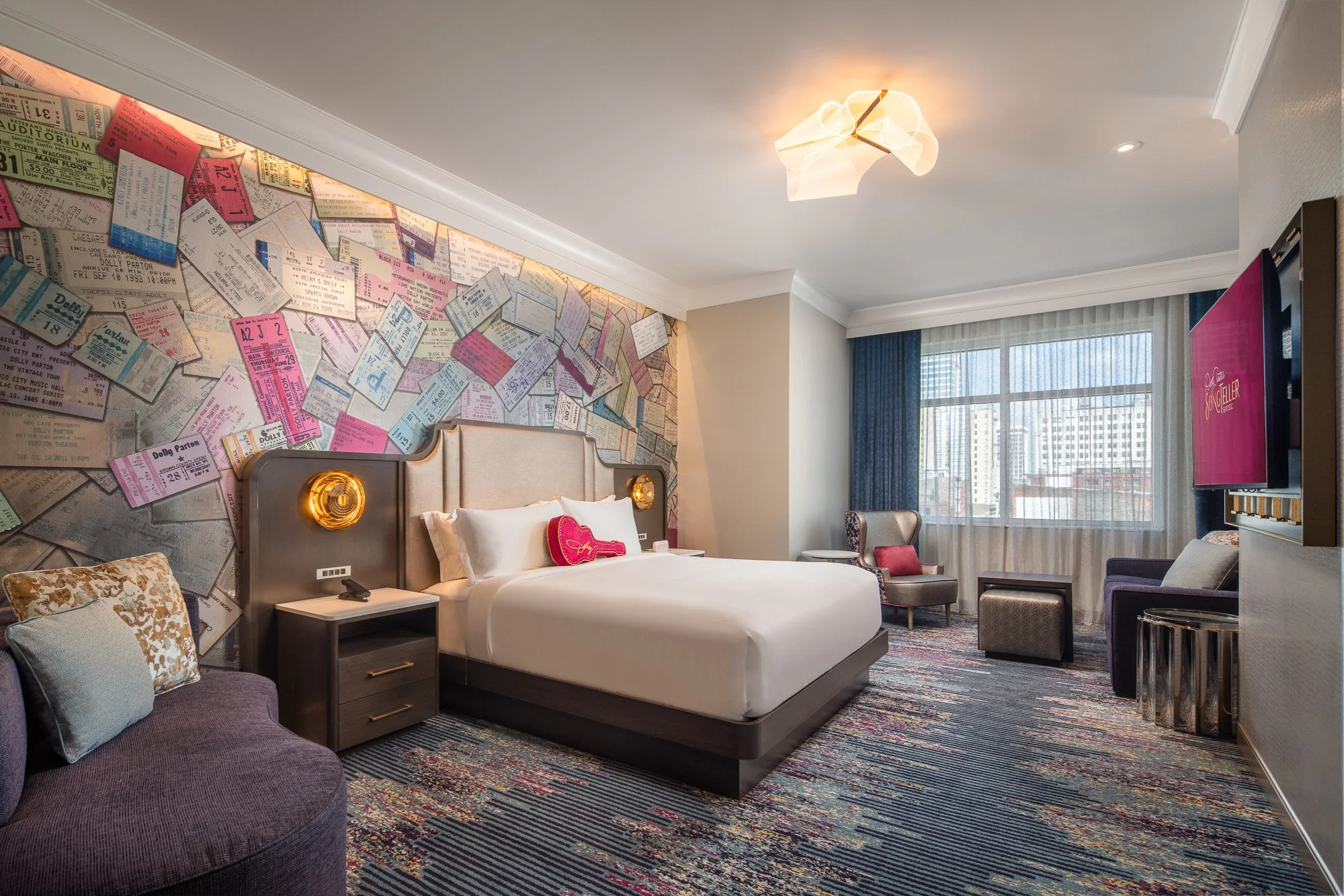 Wide view of a SongTeller Hotel guest room with a concert-ticket feature wall, upholstered bed, colorful carpet, seating area, and large windows.