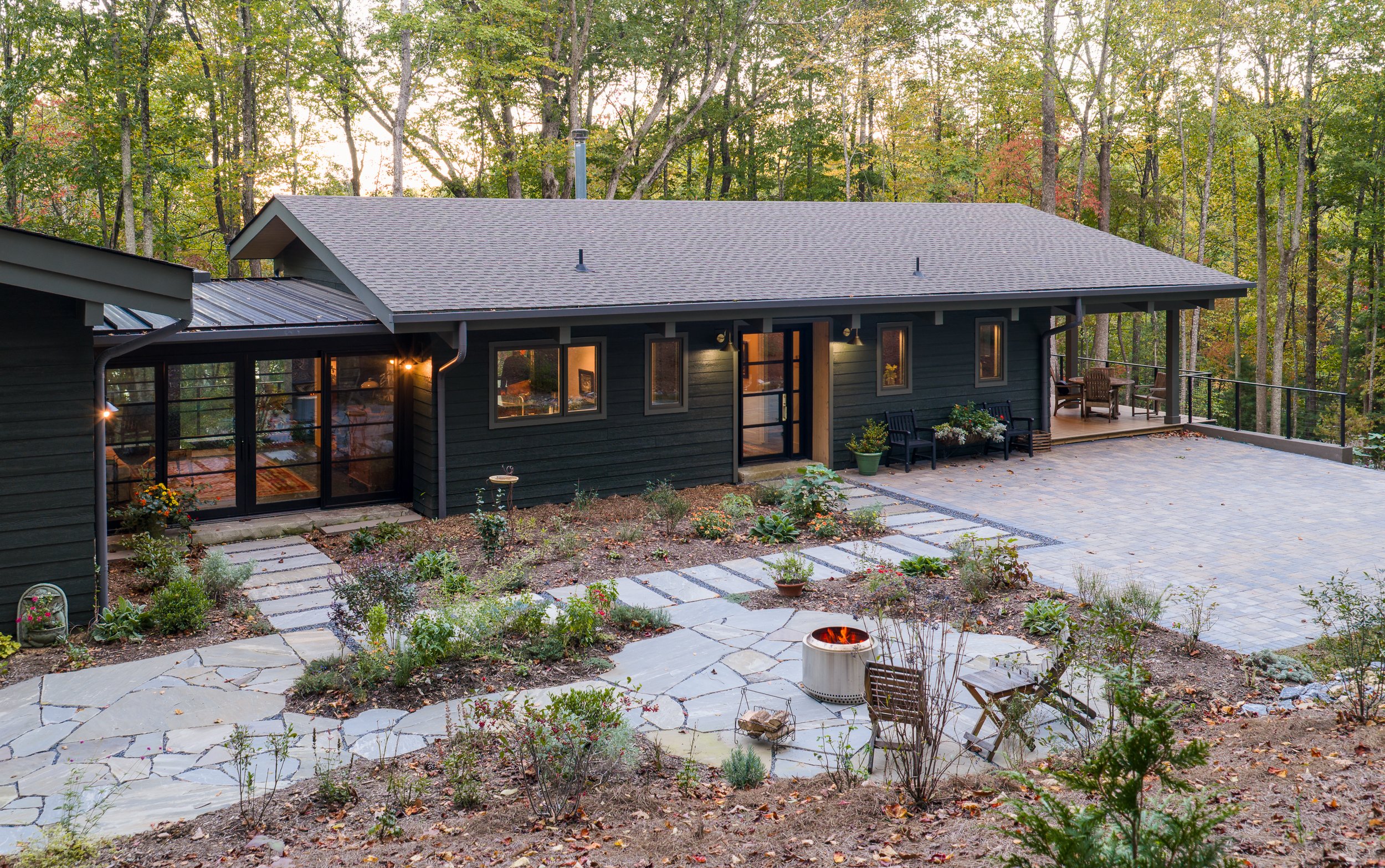 Single-story black modern cabin with stone patio, garden beds, and surrounding woods.