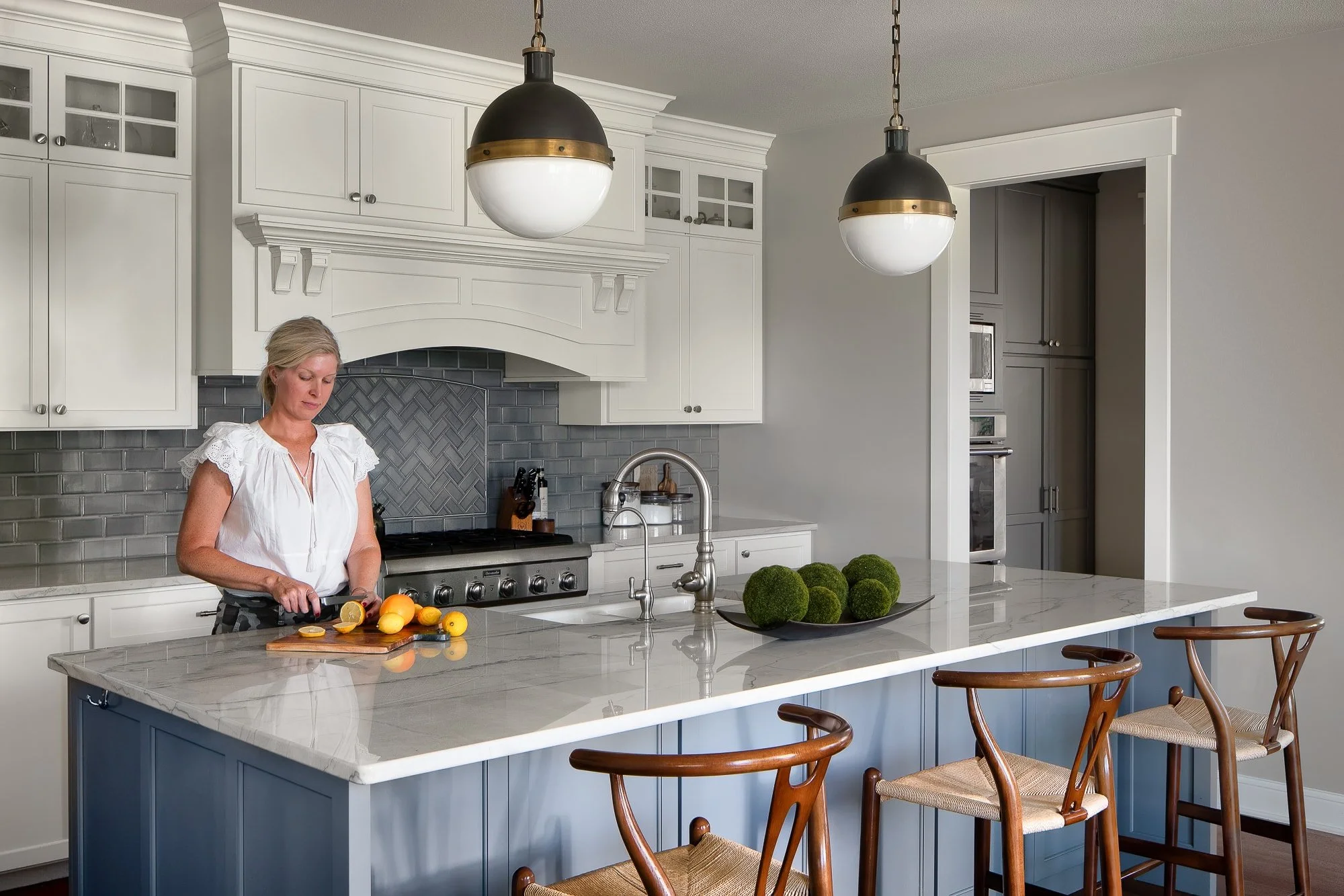Woman slicing citrus at a marble kitchen island with white cabinetry, gray tile backsplash, globe pendant lights, and woven bar stools.