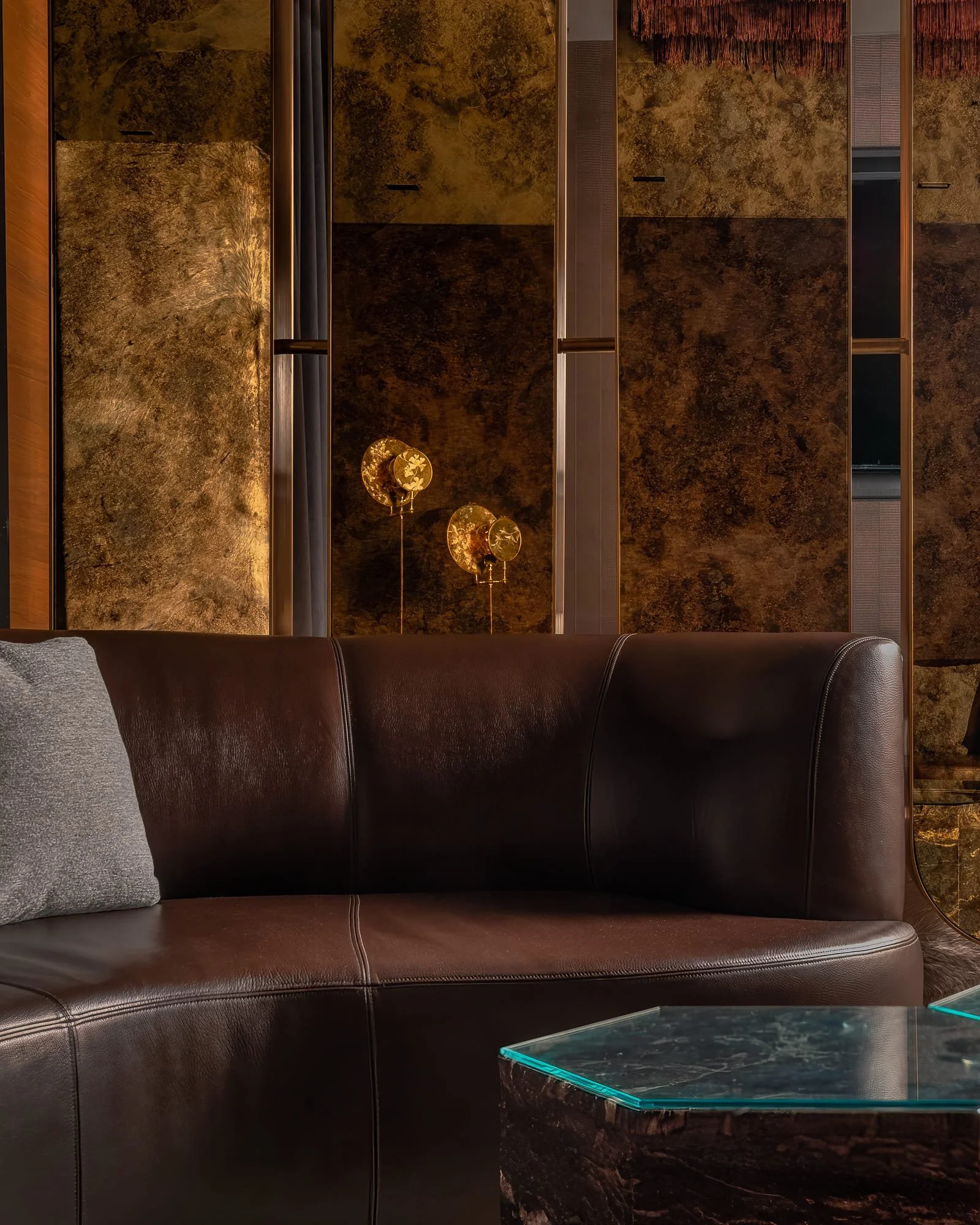 Close-up of a brown leather sofa with textured wall panels, decorative floor lamps, and a glass-topped stone table in a moody lounge setting.