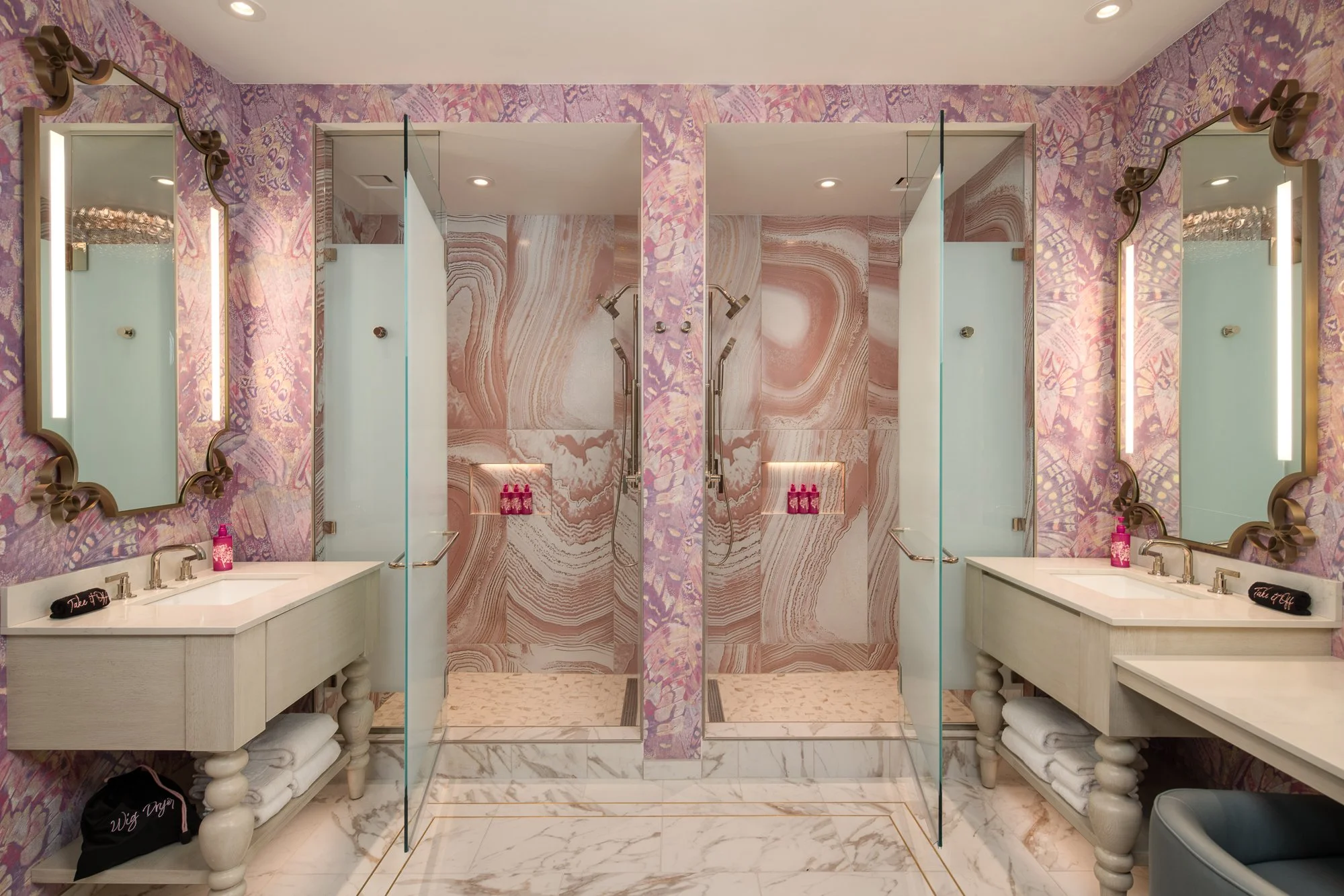 Pink and lavender hotel bathroom with two glass showers, white vanities, decorative mirrors, and dramatic stone walls.