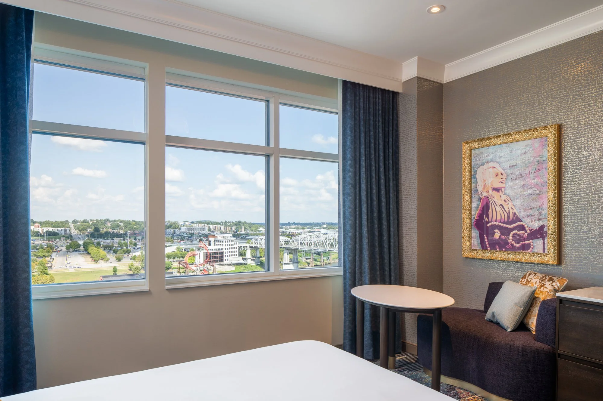 SongTeller Hotel guest room seating area beside large windows overlooking Nashville with blue drapery and framed artwork.