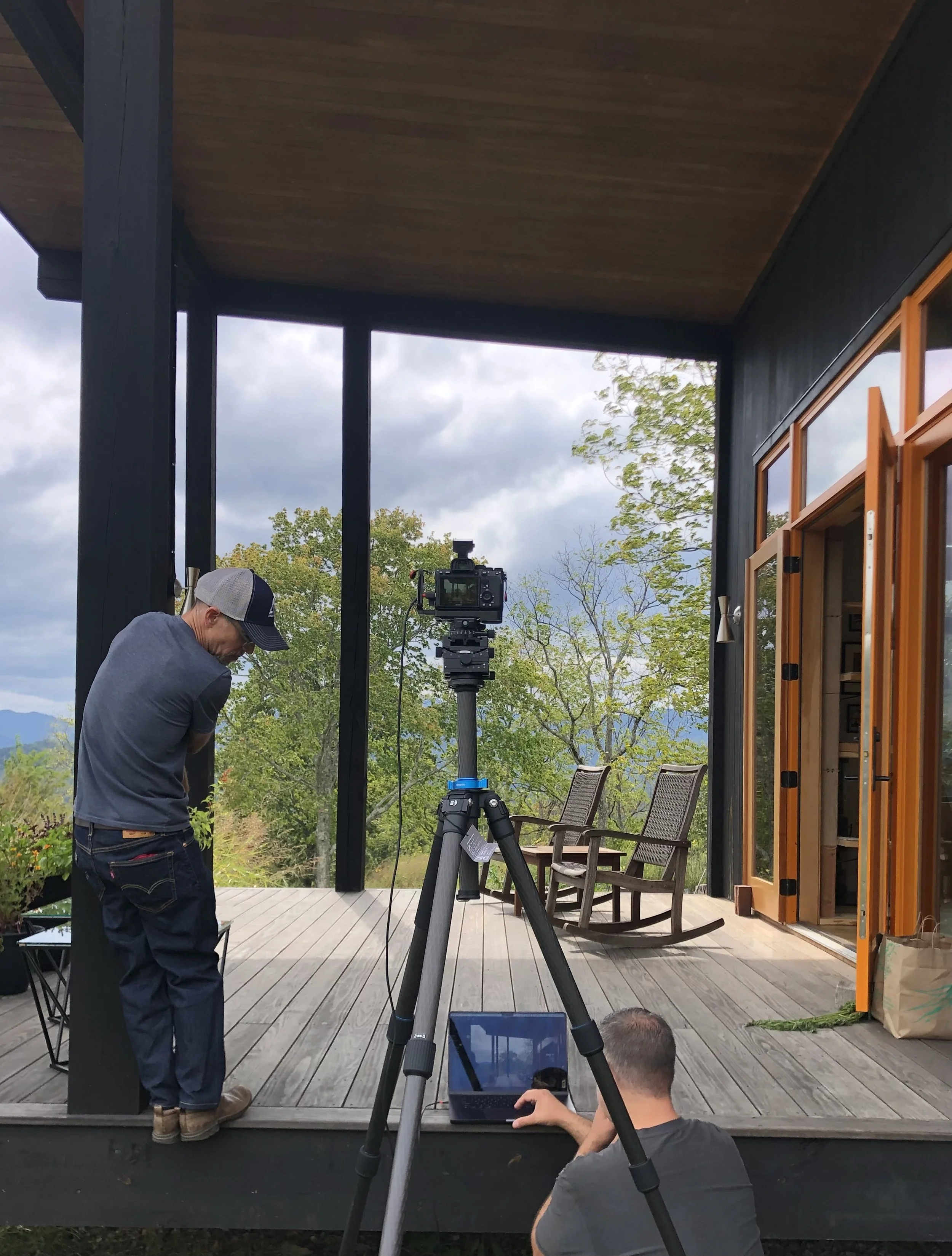 Two men setting up camera equipment on a porch with chairs, trees, and a partly cloudy sky in the background.