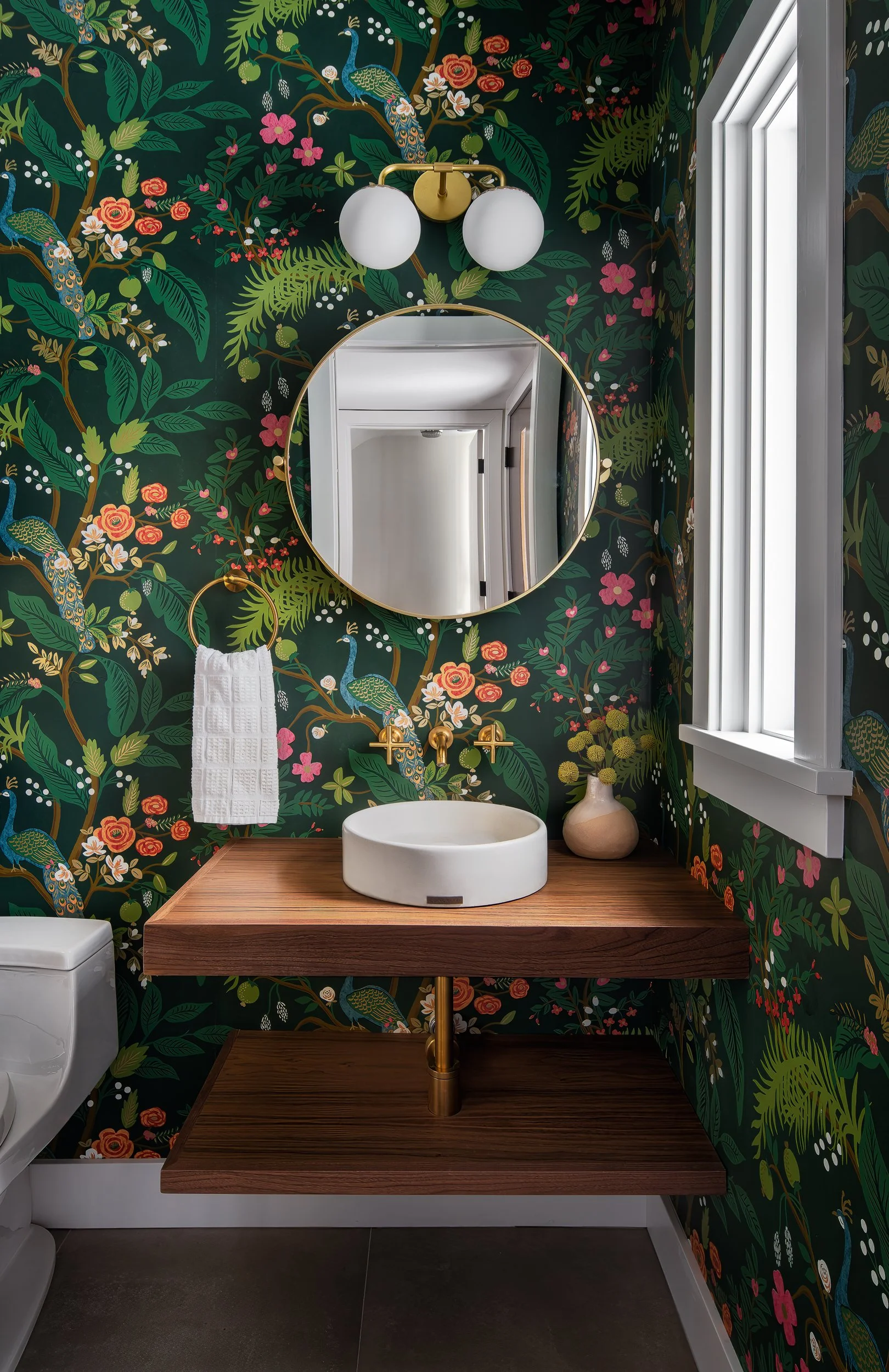 Powder room with colorful botanical wallpaper, round mirror, wood vanity, and brass fixtures.
