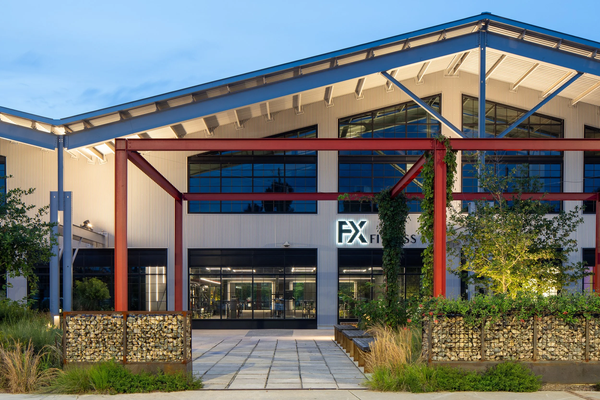 FX Fitness entrance at dusk with red structural steel framing the storefront, large industrial windows, climbing vines on the columns, and gabion stone walls flanking the paved entry path.