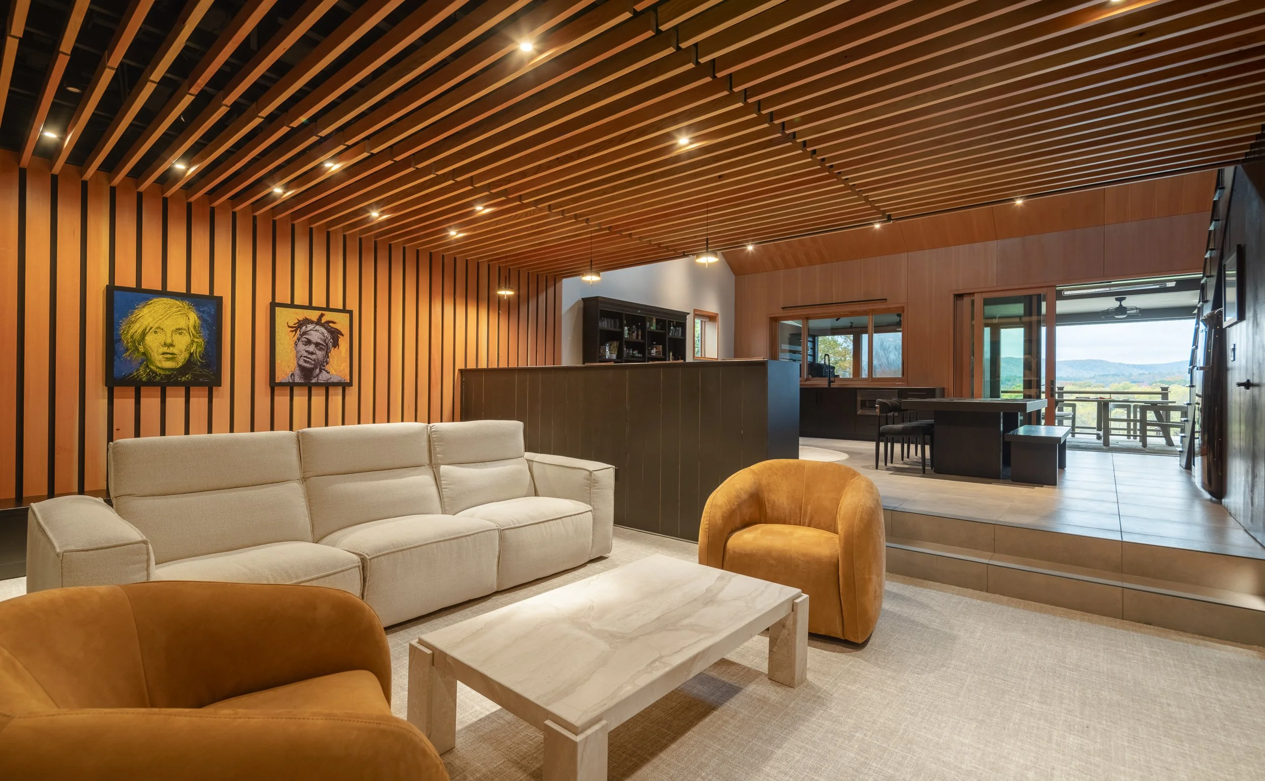 Living area with wood slat ceiling, neutral sofa, amber lounge chairs, and views beyond the kitchen.