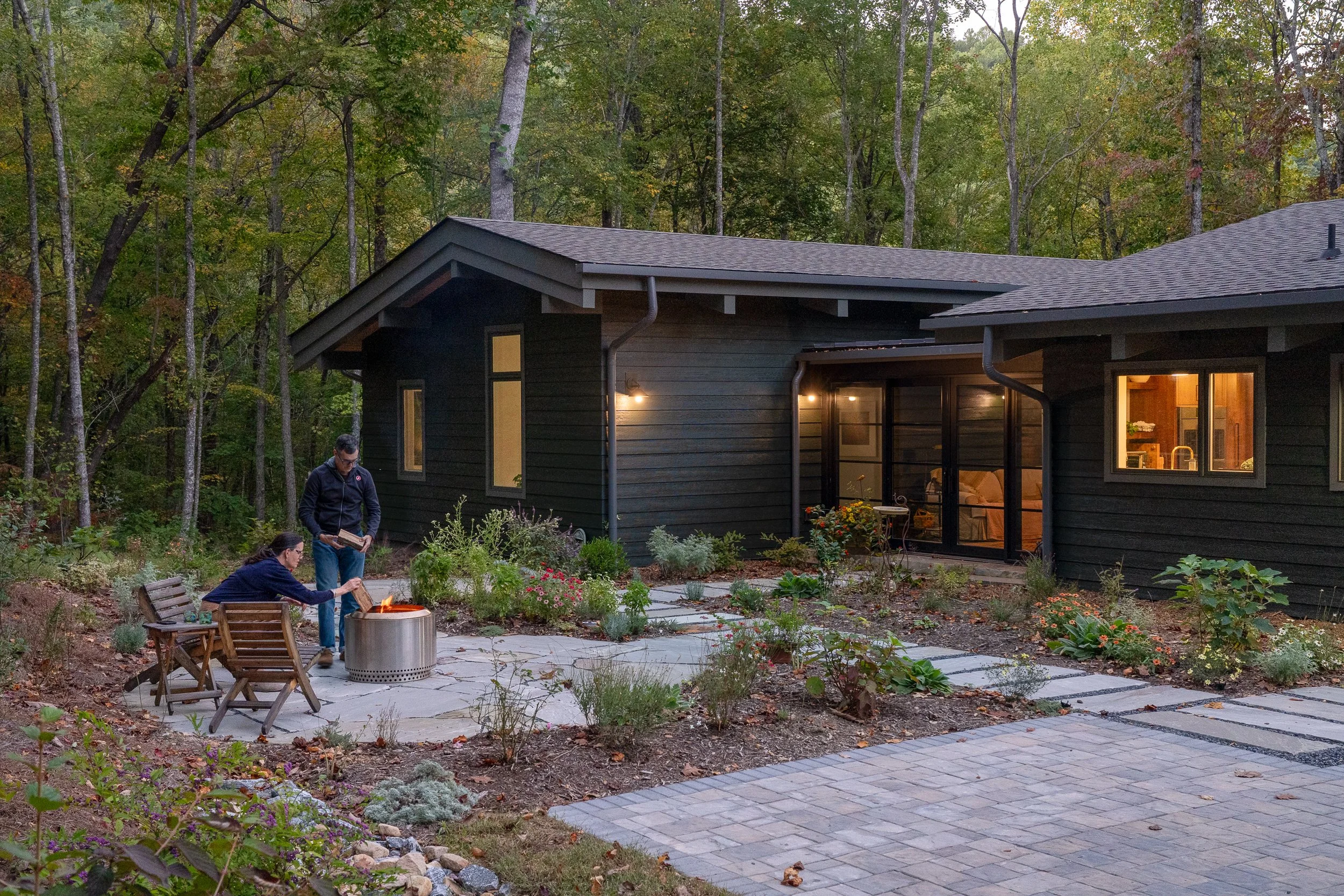 A married couple gathering around a small fire pit beside the home at dusk.