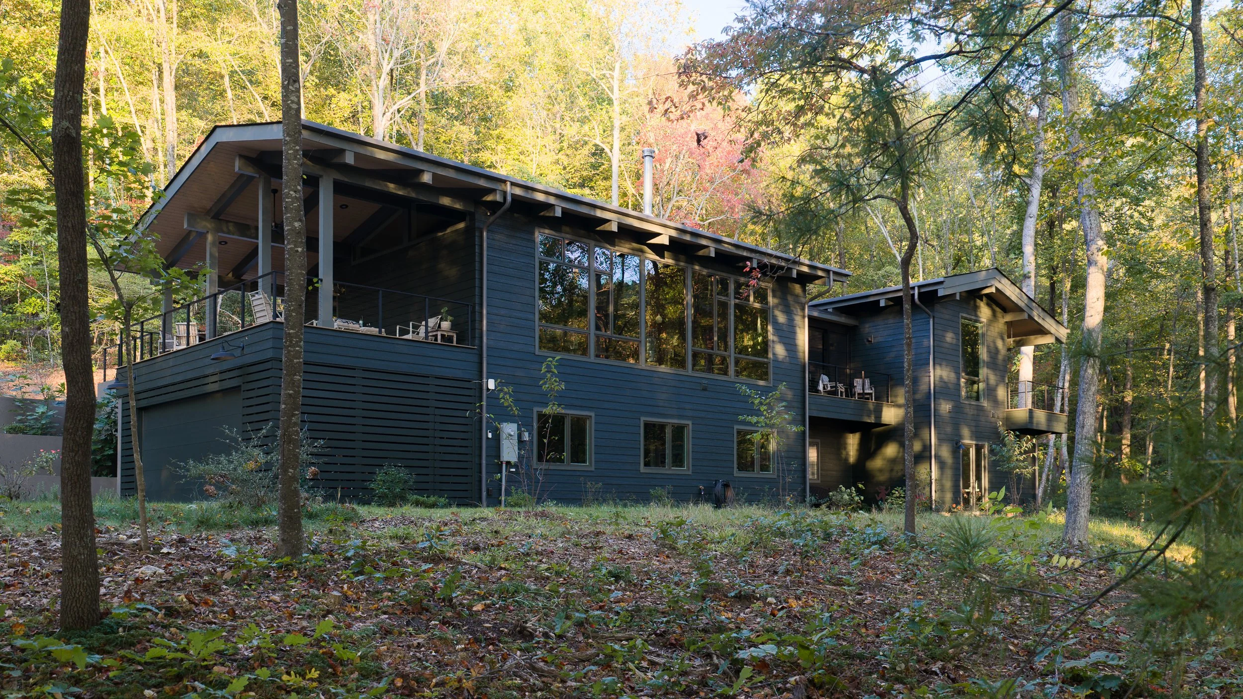 A dark-sided home sits among trees on a sloped wooded site.