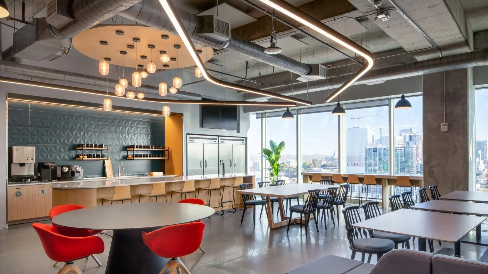 Office café with red chairs, communal tables, pendant lights, and expansive windows overlooking the city.