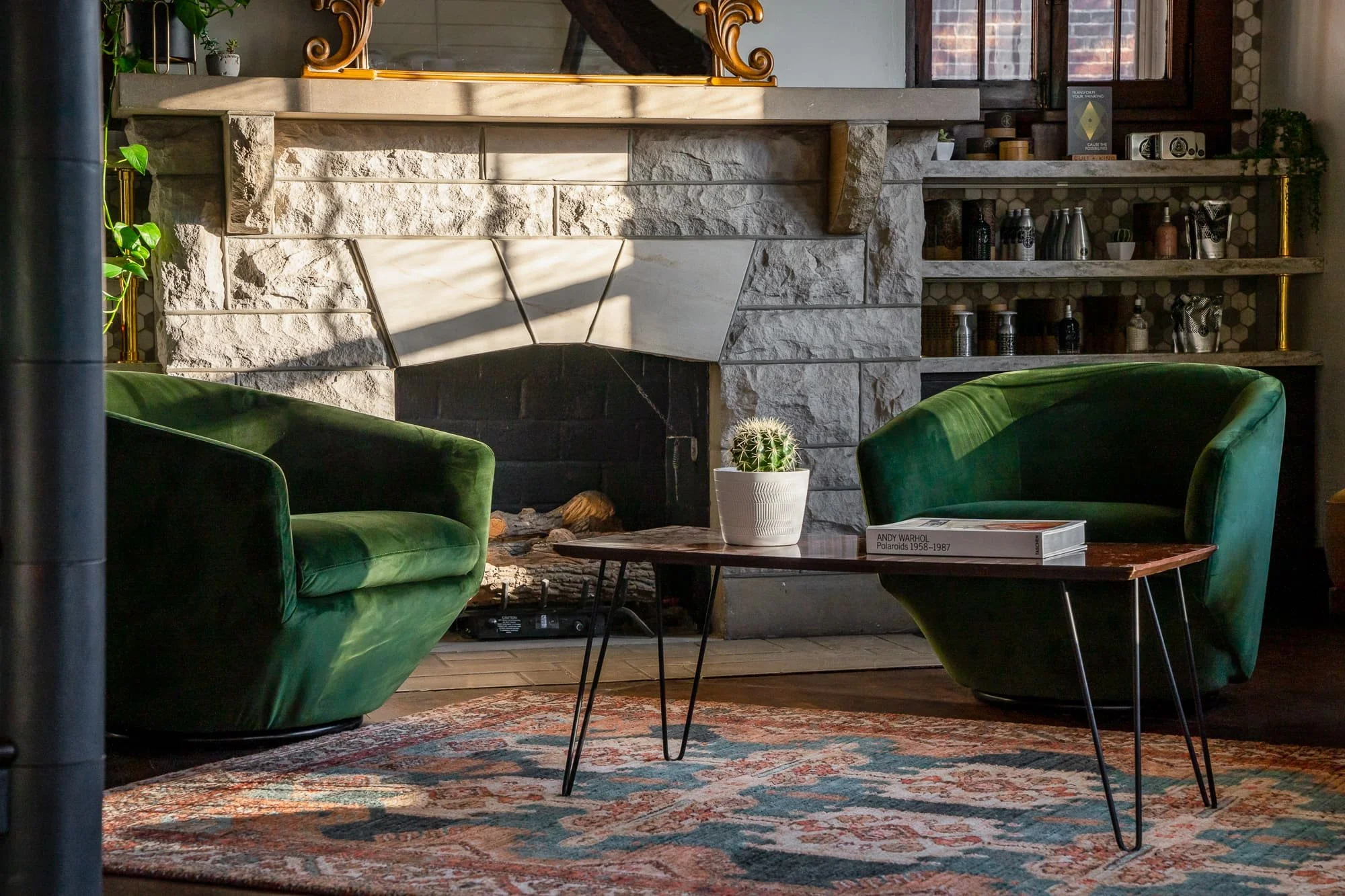 Interior lounge with green velvet chairs, stone fireplace, patterned rug, coffee table, and built-in product shelves.