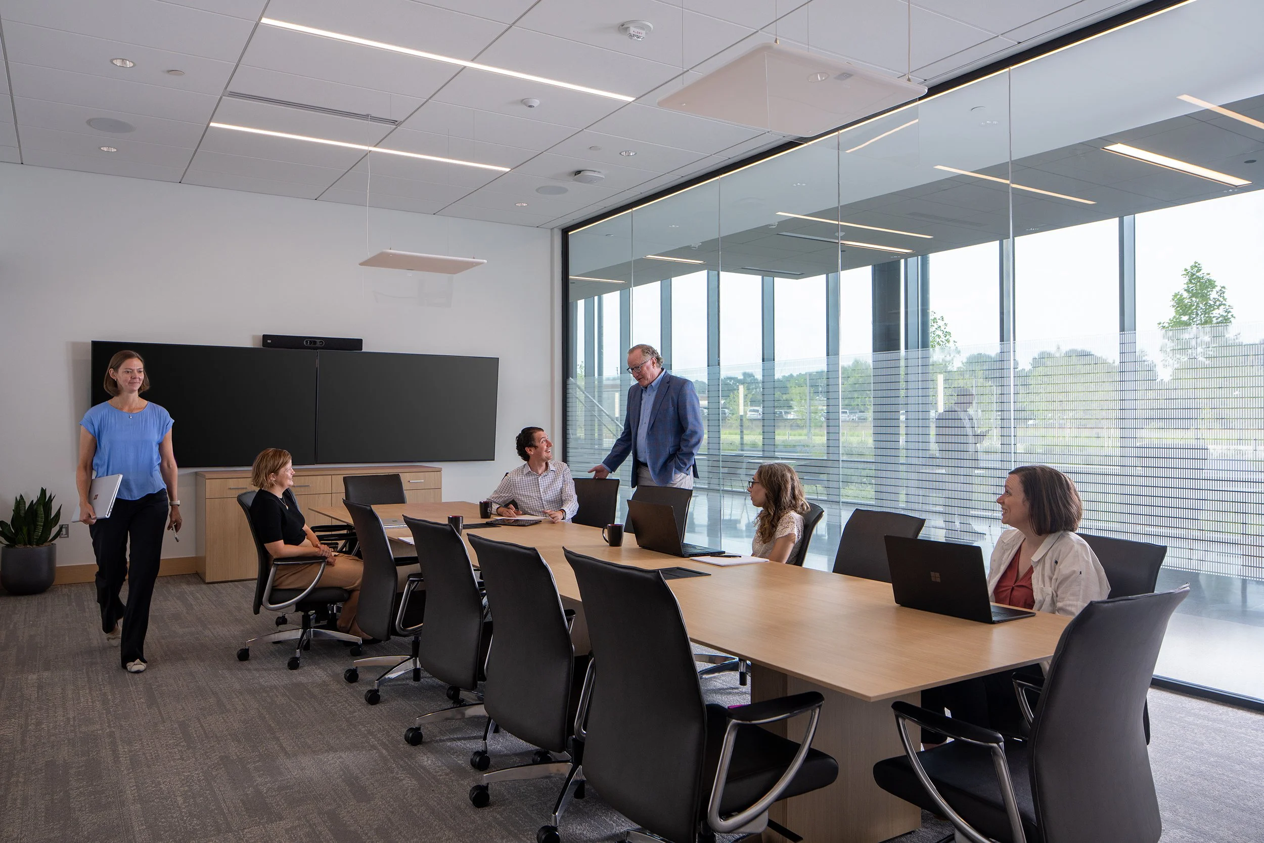 Conference room with a large video display along one wall, a long wood table, mesh chairs, and a glass partition opening to the surrounding floor with treetop views beyond.