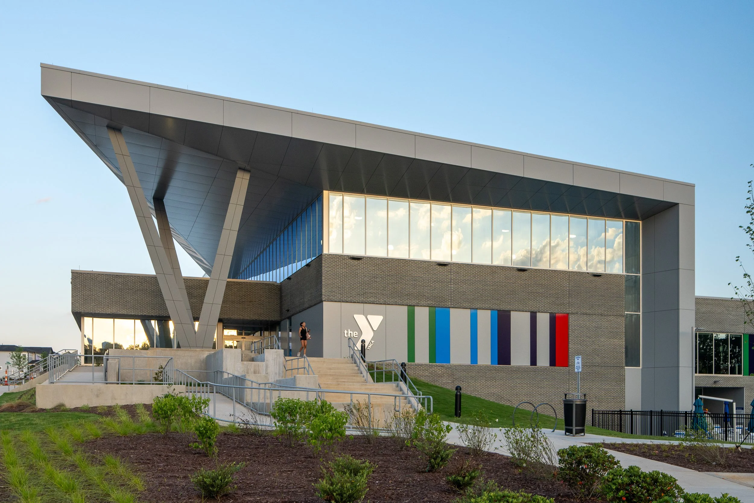 Exterior of a modern YMCA building with colorful vertical stripes on the wall, curved architecture, and a woman walking up the stairs outside.