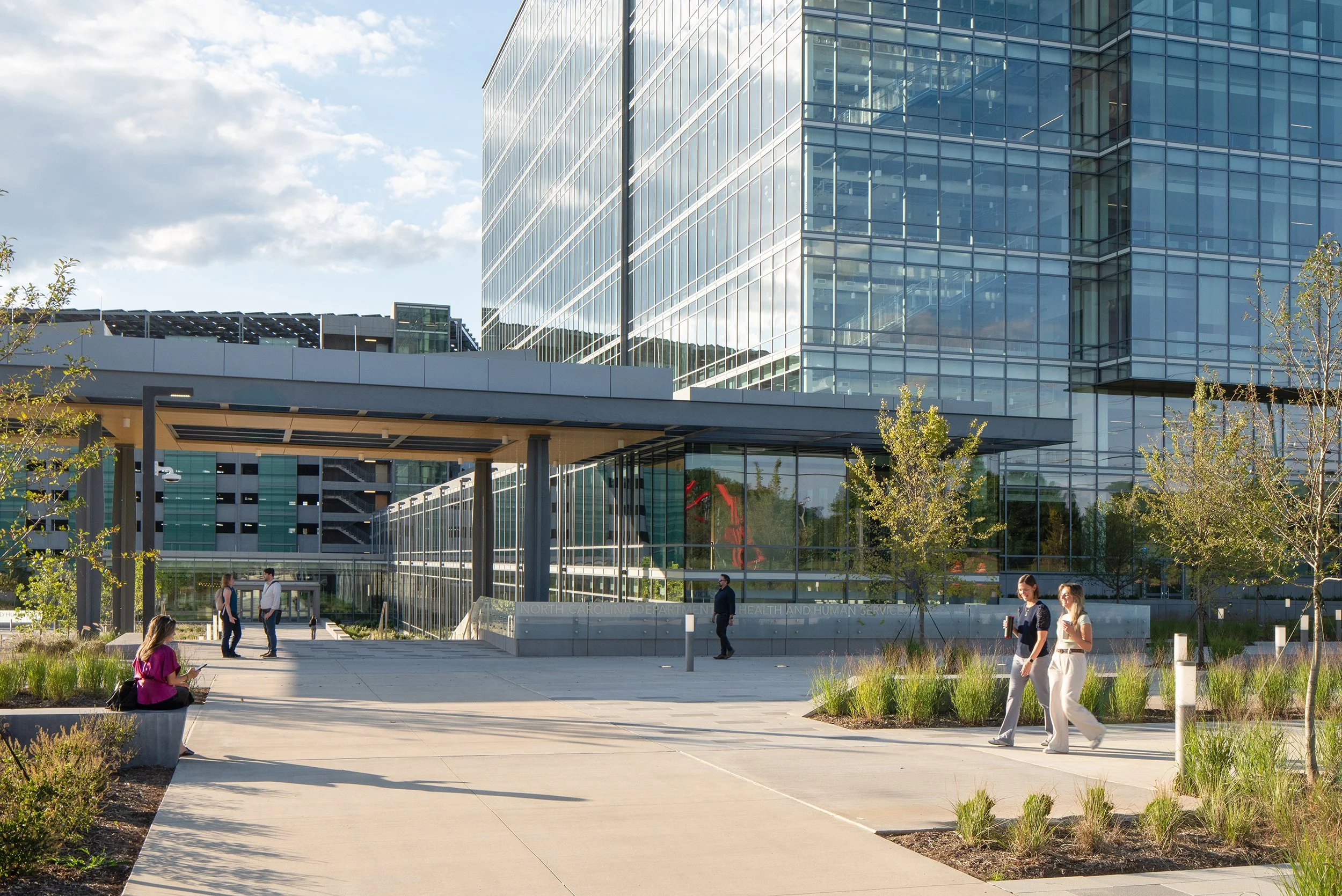 A modern glass building with people walking and sitting outside in a landscaped area.