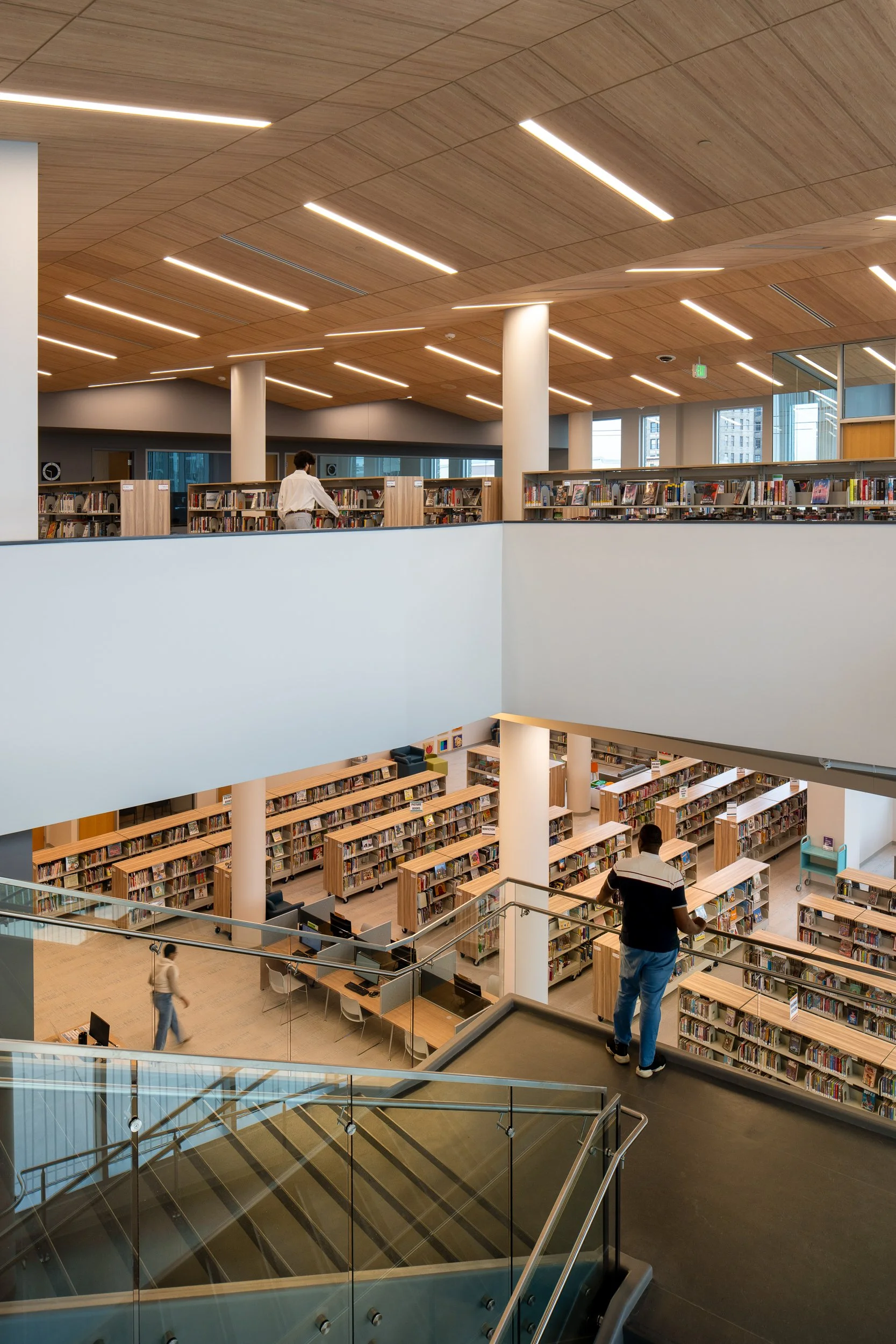 Multi-level library interior with stair, book stacks, glass railings, and warm wood ceilings.