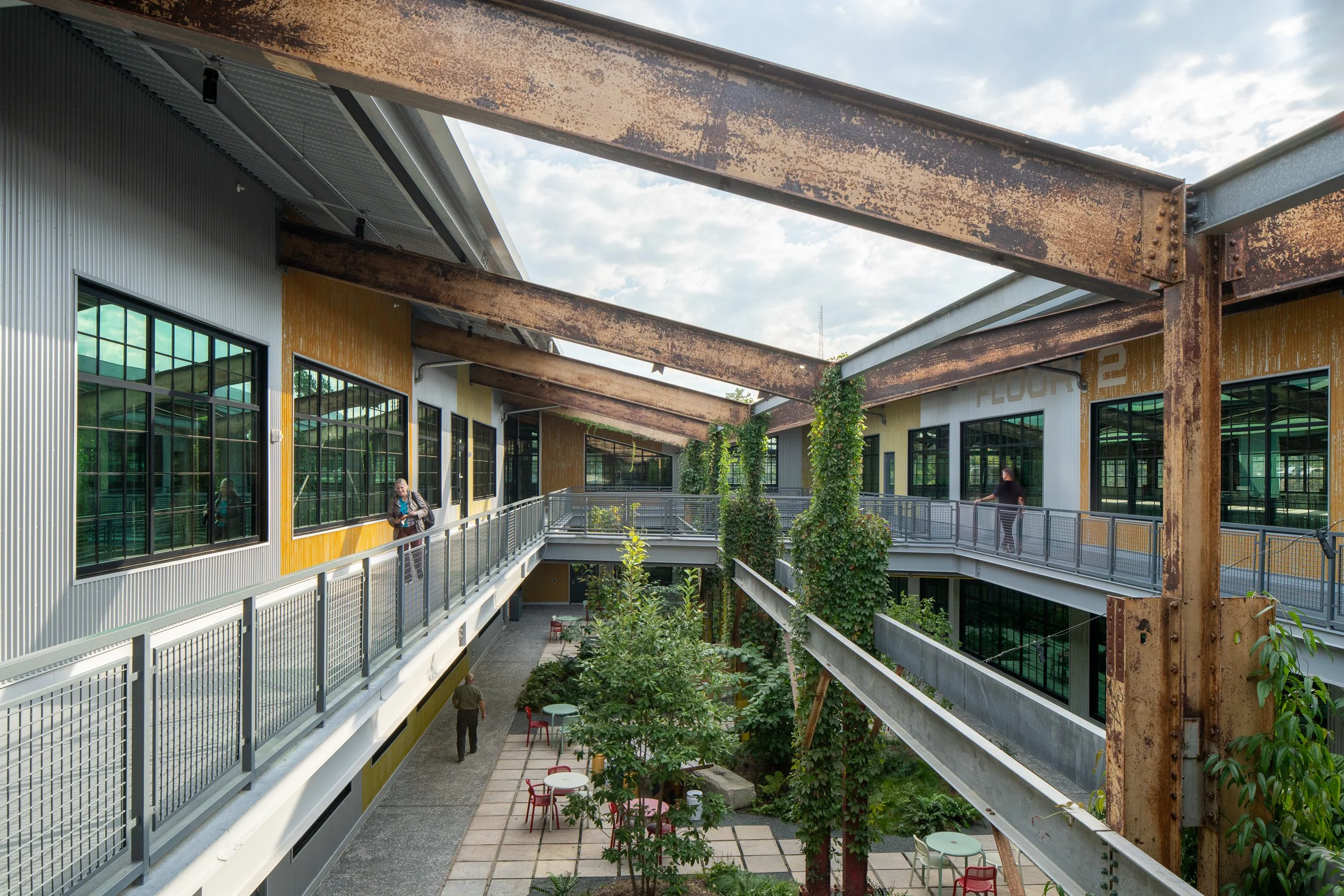 Central courtyard framed by original rusted steel trusses overhead, two levels of mesh-railed walkways, lush green plantings, and a faded FLOOR 2 ghost sign on the yellow building wall.