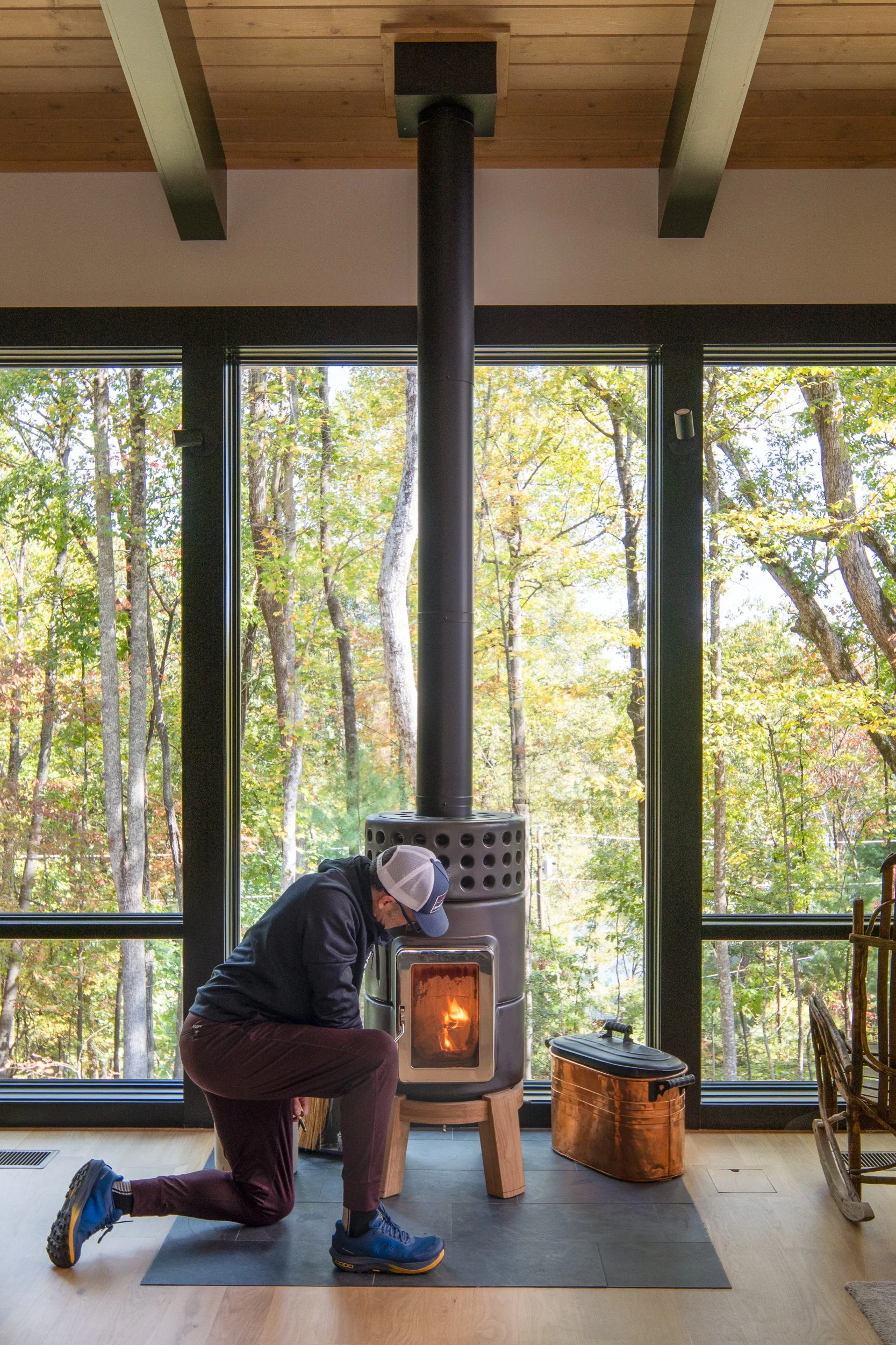 A man kneeling to tend the wood stove beside large windows.