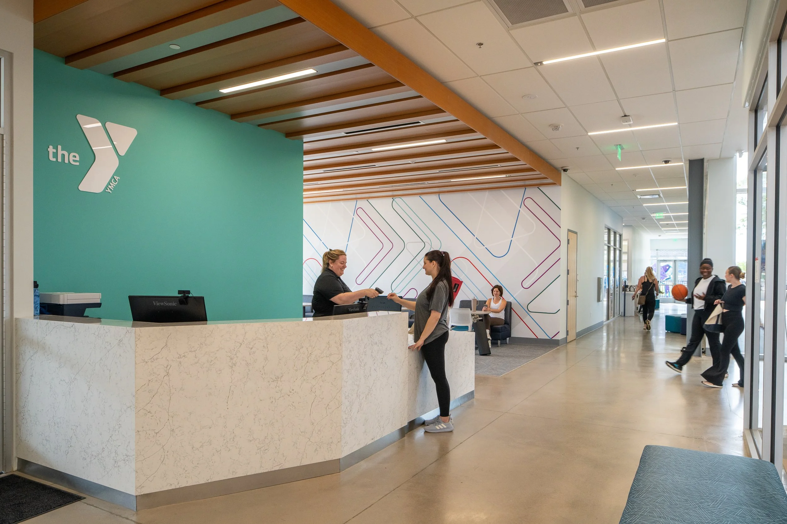 YMCA lobby with teal brand wall, marble-look reception desk, wood ceiling slats and members checking in.