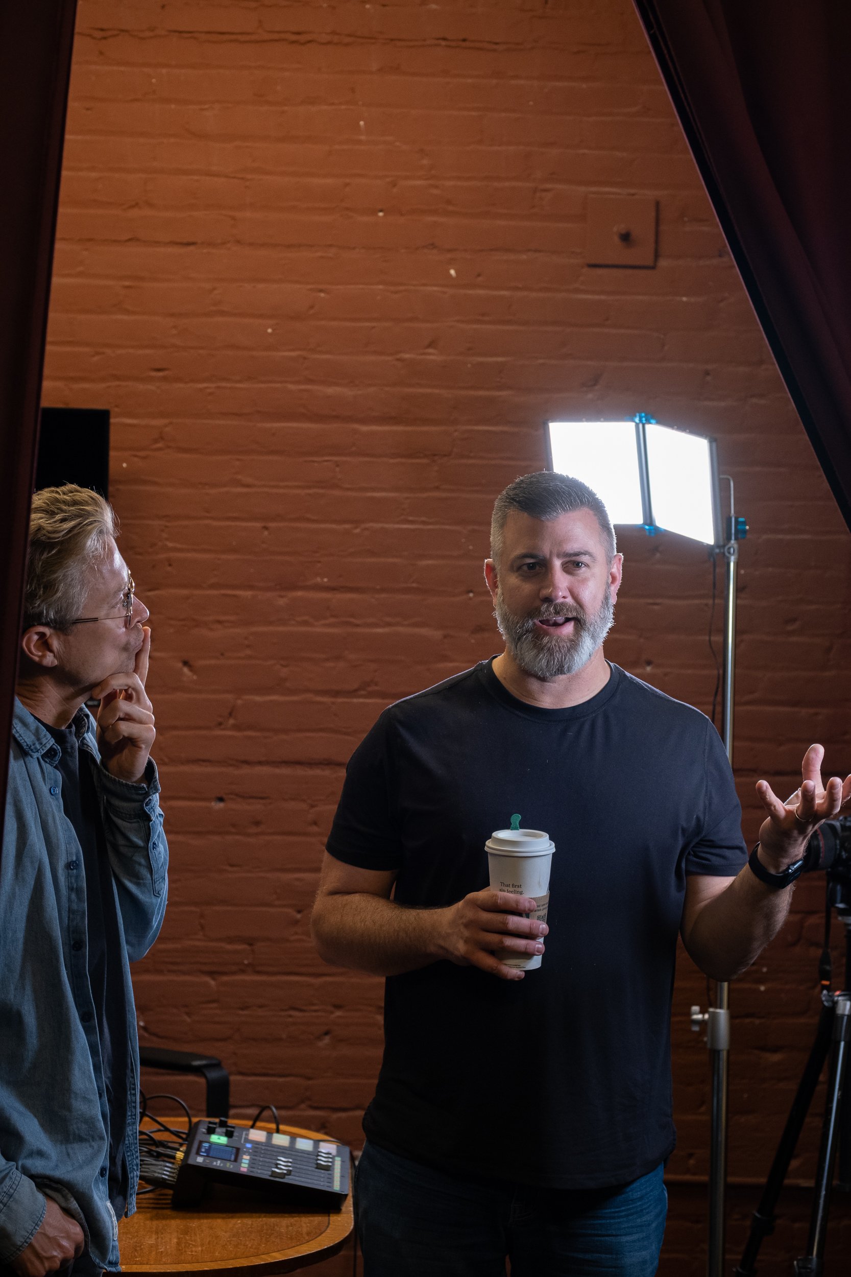 A man with a beard and short hair, wearing a black t-shirt, is holding a coffee cup and speaking, with a woman with glasses and gray hair sitting nearby, and studio lighting and audio equipment visible in the background.