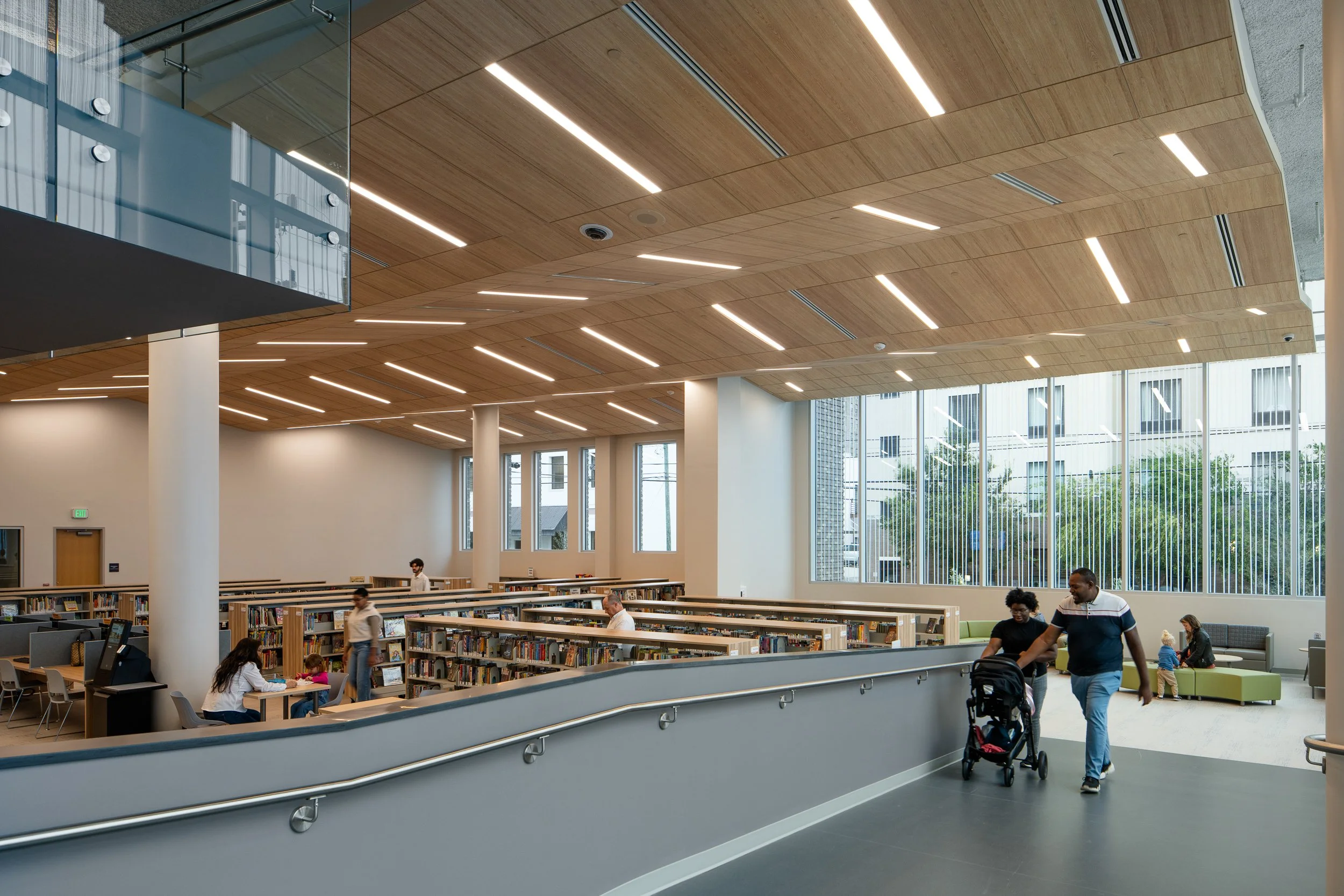 Library interior with curving ramp, book stacks, wood ceiling panels, tall windows, and visitors.