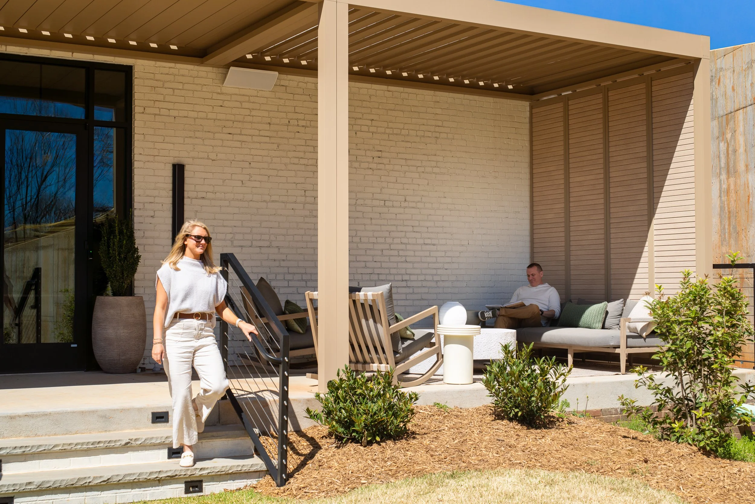 Rear patio with a louvered metal pergola sheltering a cushioned lounge sofa and rocking chairs, painted brick exterior wall, cable-rail steps leading down, and staff using the outdoor space.