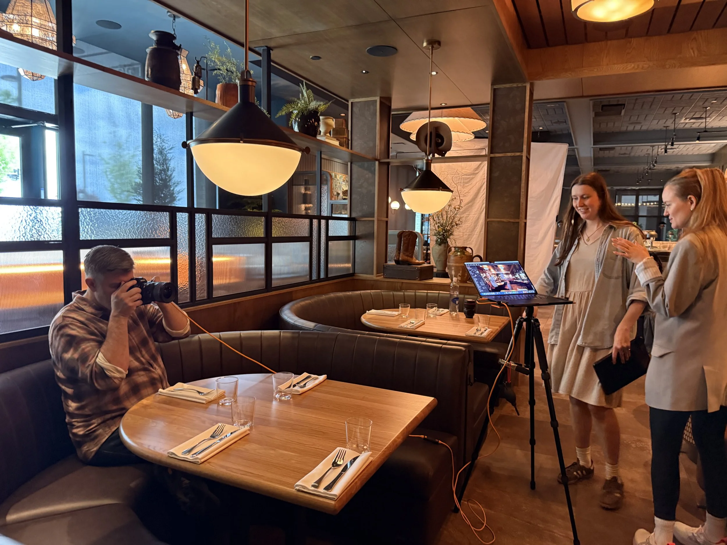 Two women on set preparing for a photo shoot in a restaurant with warm lighting, wooden decor, and large windows. One woman is looking at her phone, while the other is smiling and looking down. A man with glasses is sitting at a table with a camera, 