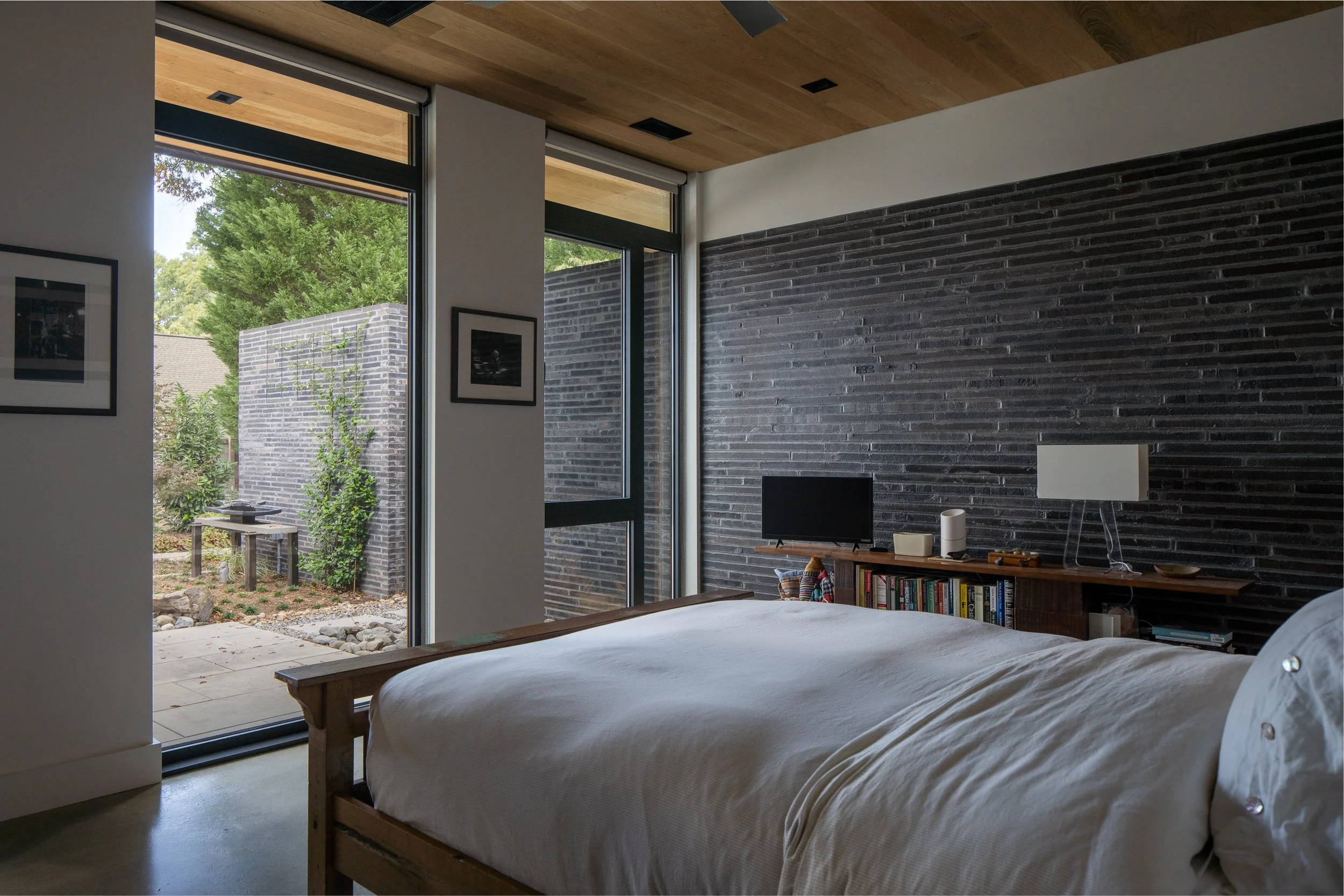 A bed faces full-height windows overlooking an enclosed outdoor area, with a dark brick wall and wood ceiling defining the bedroom.