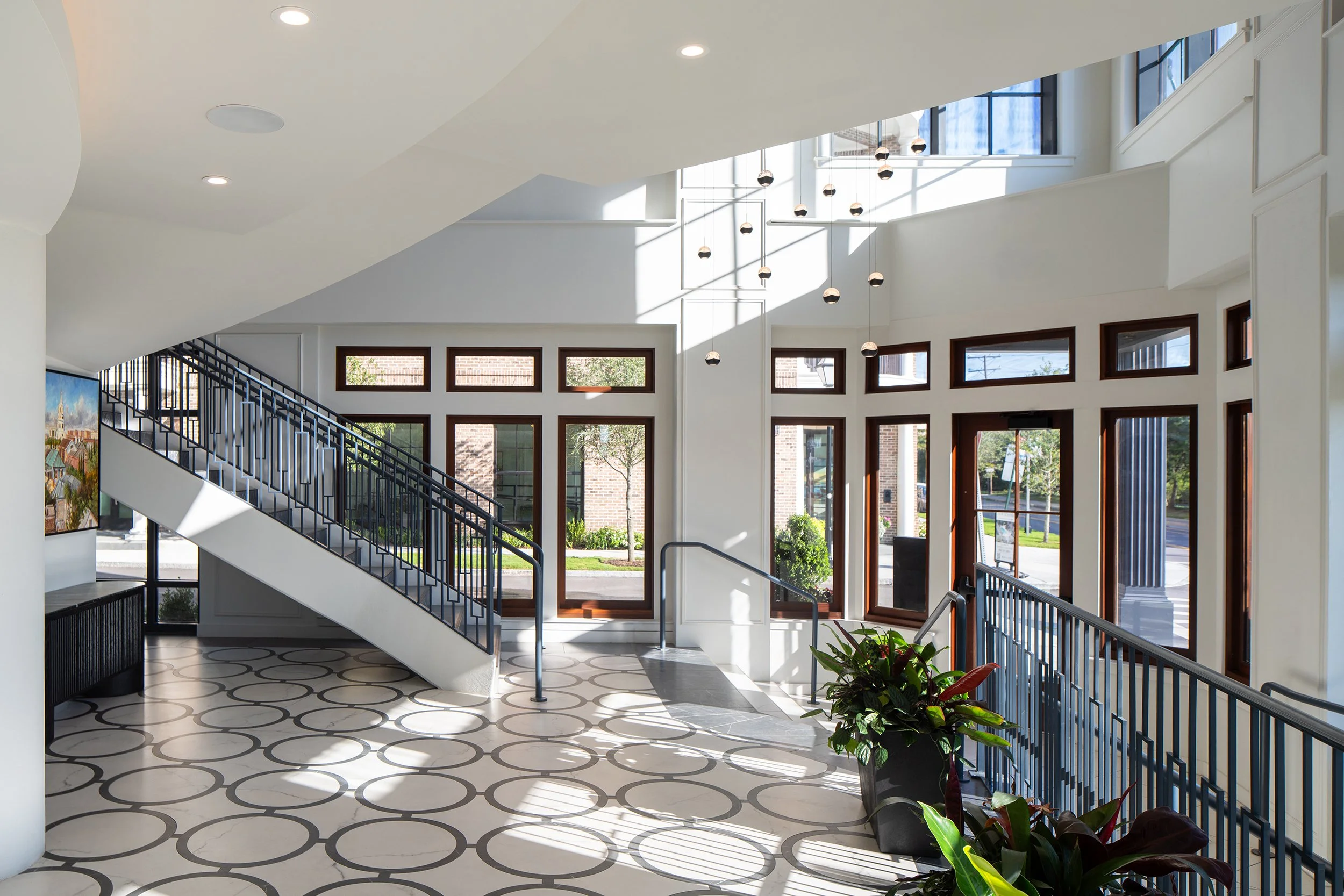 Lobby interior looking back toward the street entry with the dark marble stair on the left, the geometric ring-pattern marble floor, double-height windows with mahogany trim, and a constellation of pendant globe lights overhead.