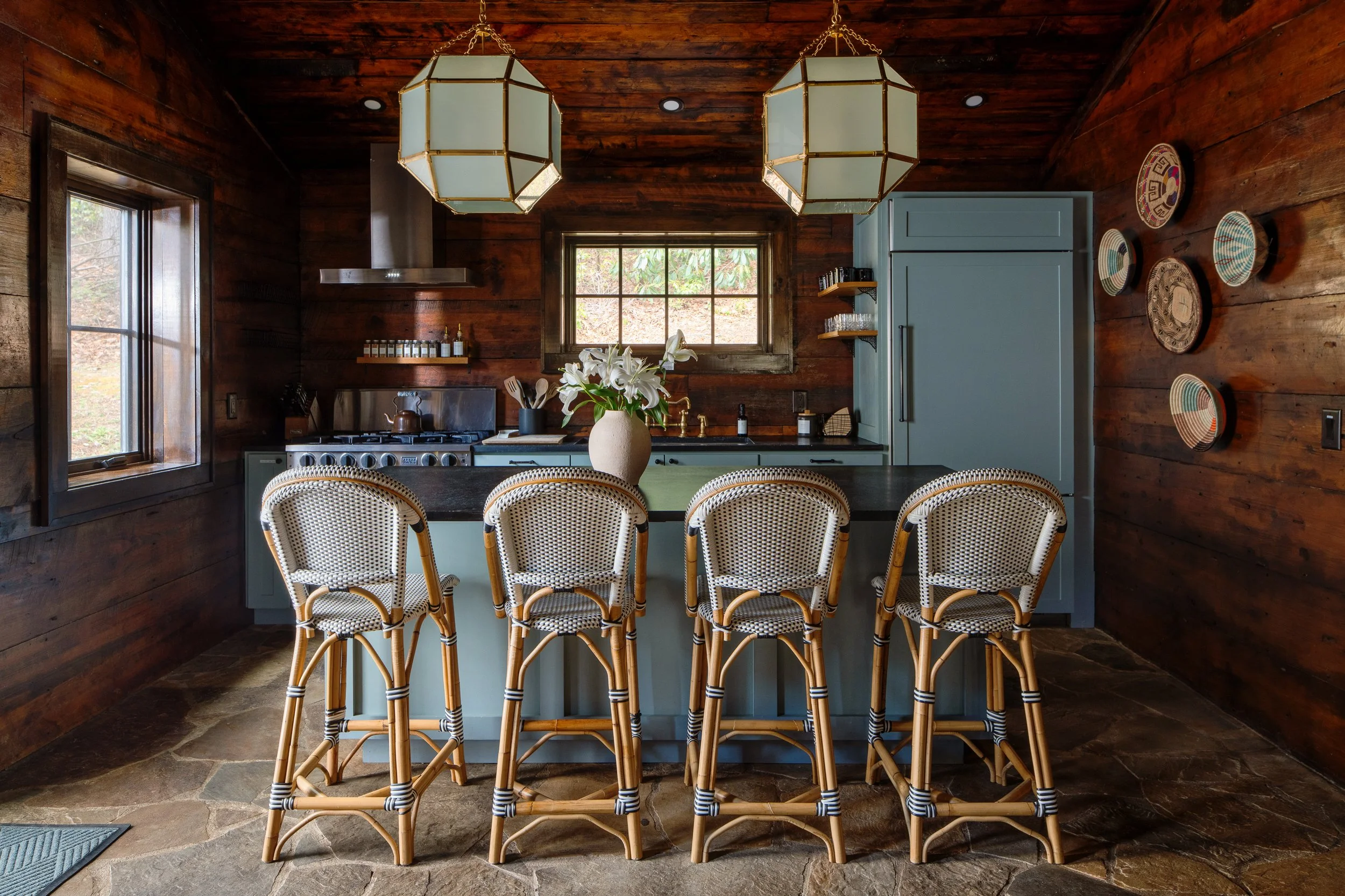 Rustic cabin kitchen with wood-paneled walls, blue island, bistro stools, stone floor, and two pendant lights.