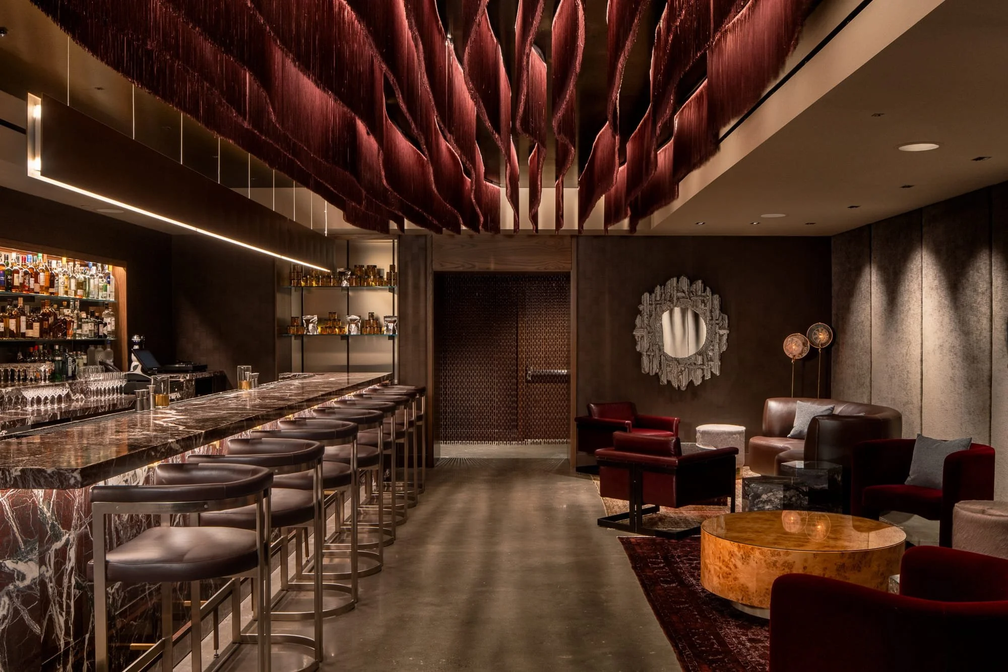 Interior of a bar or lounge with a marble counter, bar stools, and a variety of bottles on shelves. There are comfortable chairs and tables, an ornate mirror, and warm lighting with a modern decorative ceiling feature.