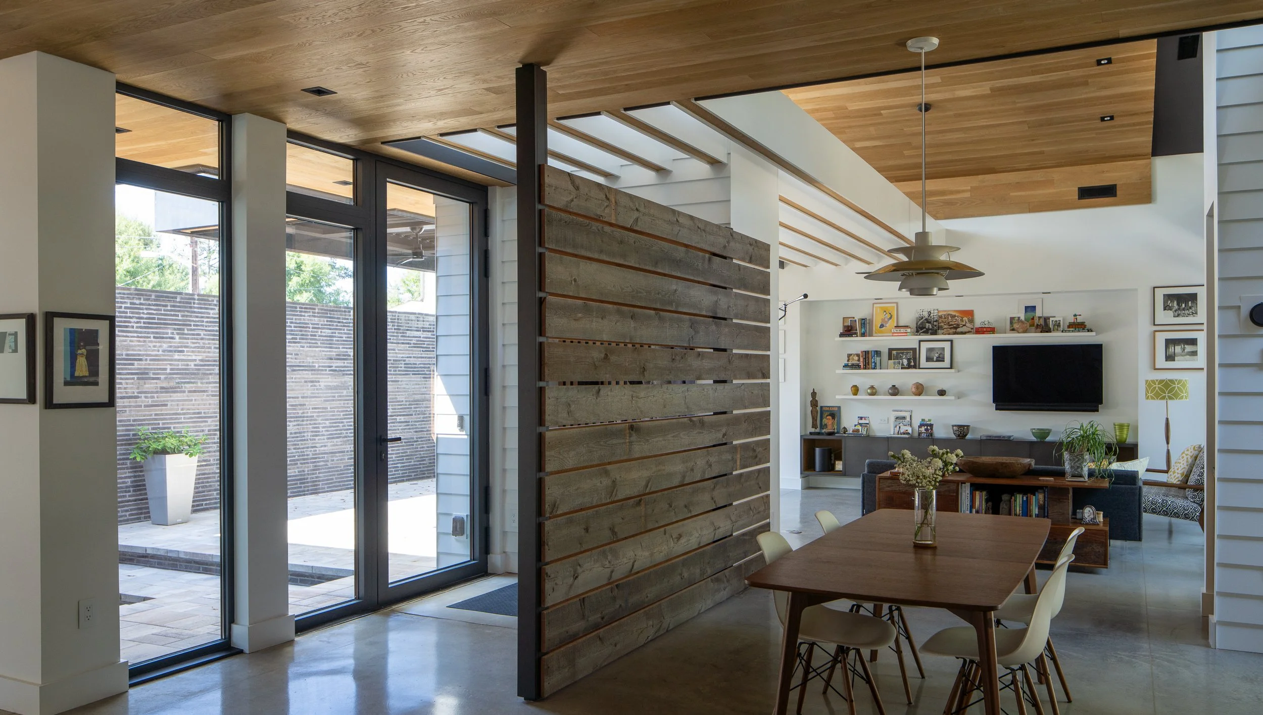 A wood dining table sits beside courtyard glazing and a horizontal wood partition, with the living room visible beyond.