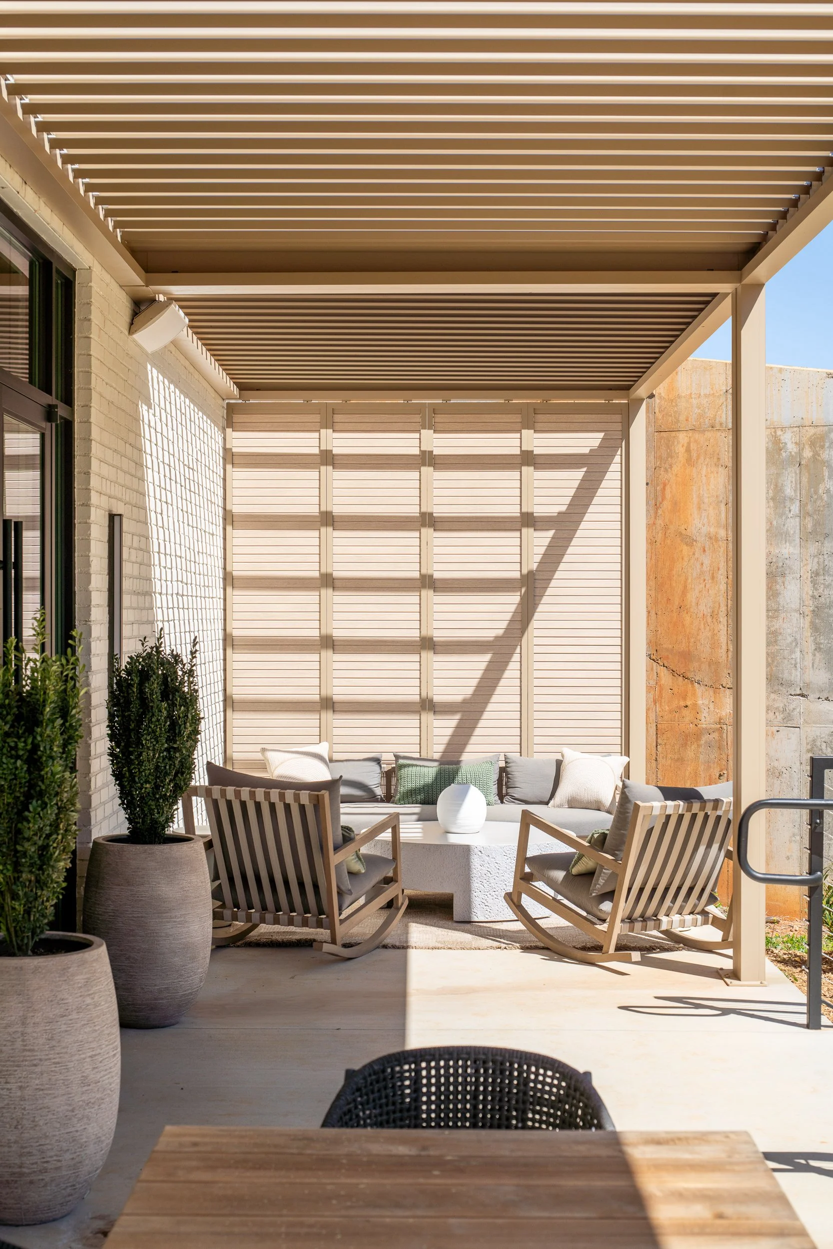 Outdoor covered patio with a louvered metal pergola overhead, two wood rocking chairs, a low concrete coffee table, slatted privacy screening, and tall potted topiaries casting striped shadows.