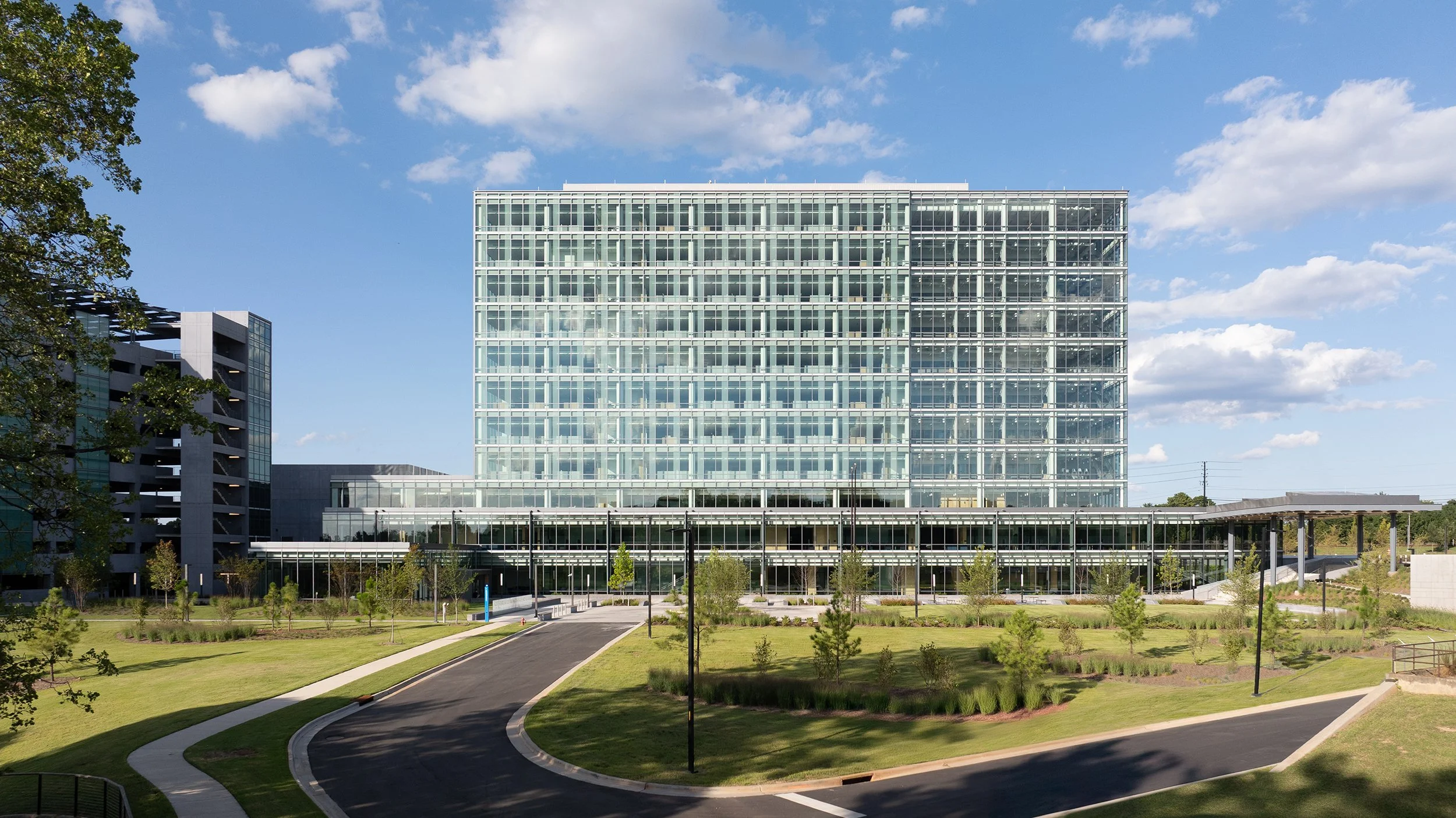 Wide daytime view of the campus showing the glass office tower, adjacent parking deck, lower glass concourse, and curved drive lined with native grasses.