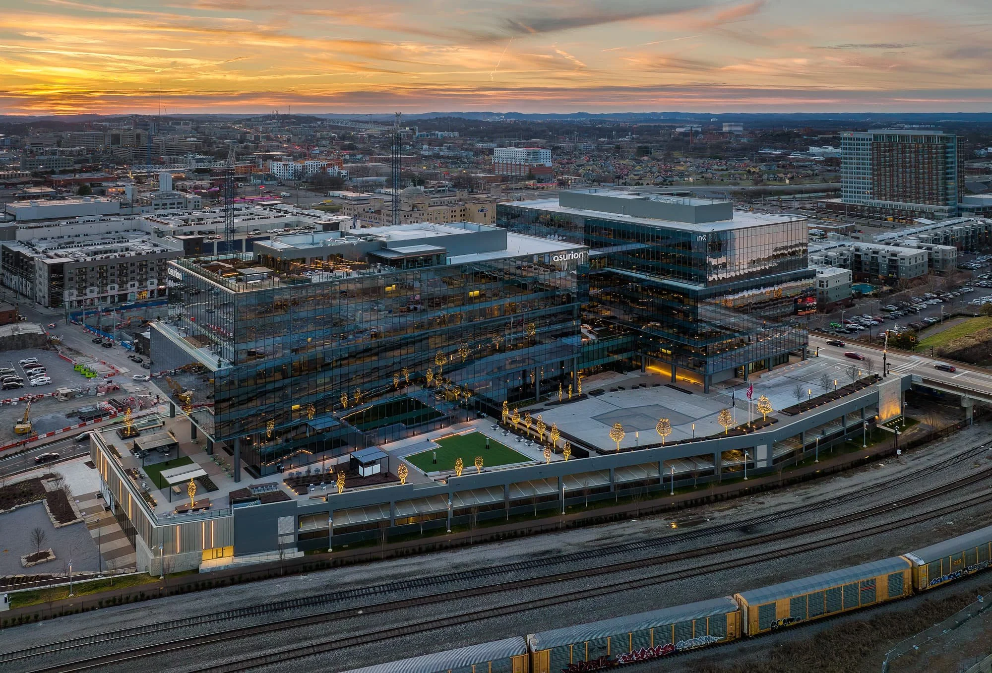 Aerial drone view of the Asurion headquarters campus in Nashville Yards at sunset with terraced rooftop amenities, illuminated trees, freight rail line below, and a dramatic orange sky