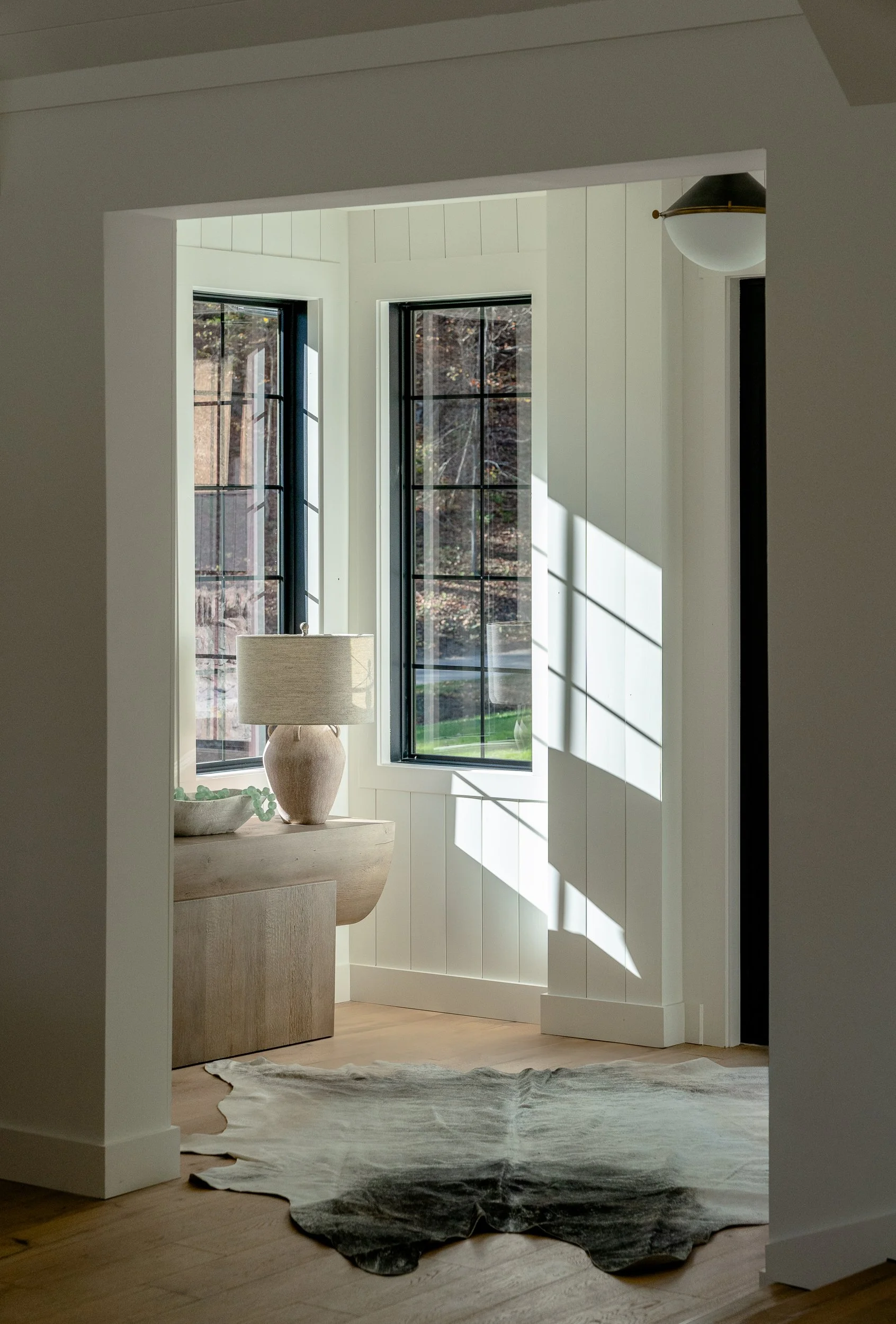 Sunlit interior entry with black-framed windows, white paneled walls, a console table, lamp, and hide rug.