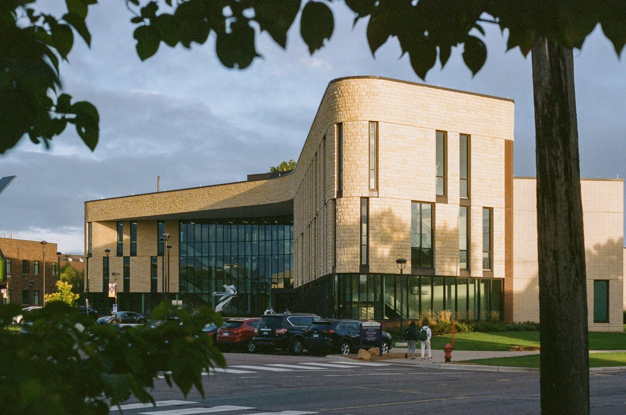 A modern building with a curved design, made of light-colored bricks and large glass windows, surrounded by a parking lot with several cars, trees, and a few people walking nearby. The sky is partly cloudy with some sunlight.
