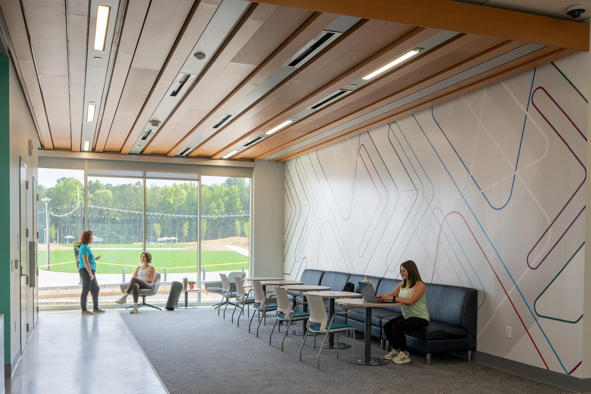 Community lounge with café seating, patterned wall graphics and large windows looking toward outdoor fields.