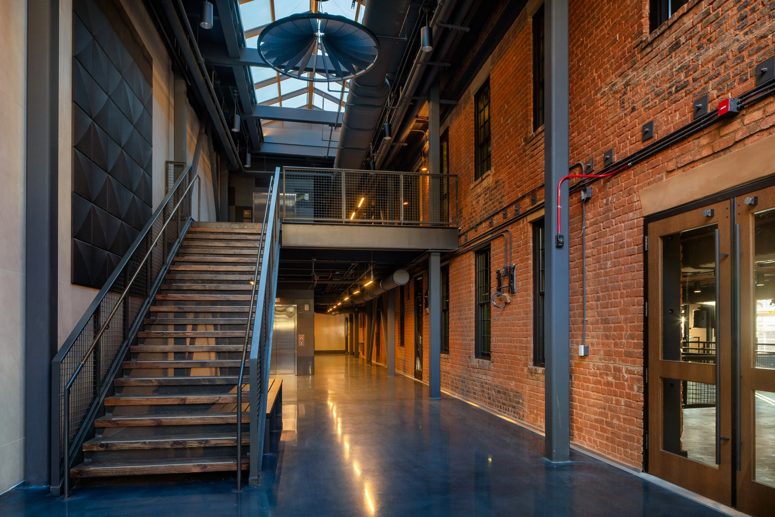 Skylit concourse with a steel and wood stair on the left, an original brick wall with steel-framed windows on the right, and polished concrete floors running the length of the space.