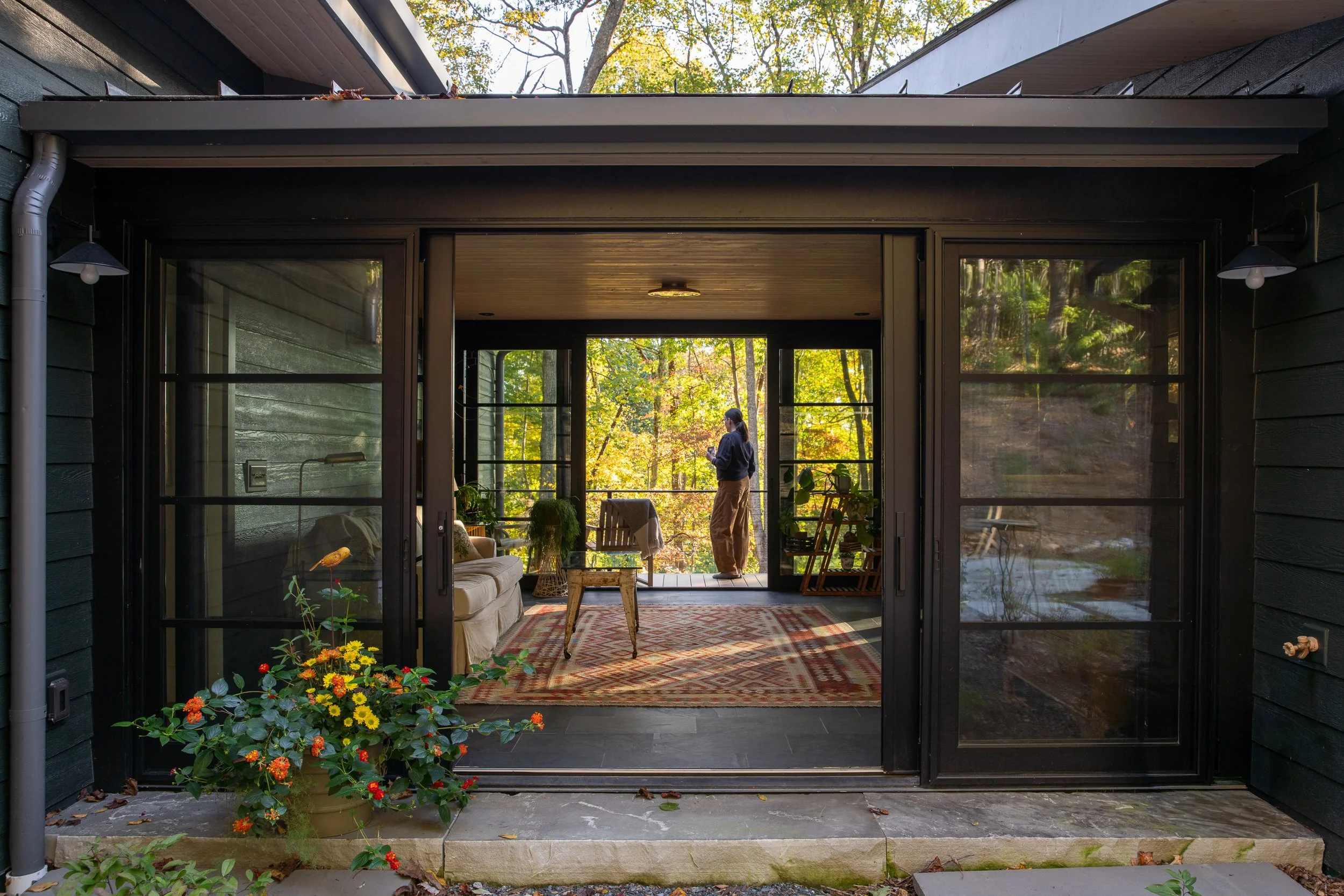 A person standing on the porch of a house surrounded by trees, with large sliding glass doors opening into a cozy interior room decorated with plants, a sofa, and a colorful rug, during daytime.