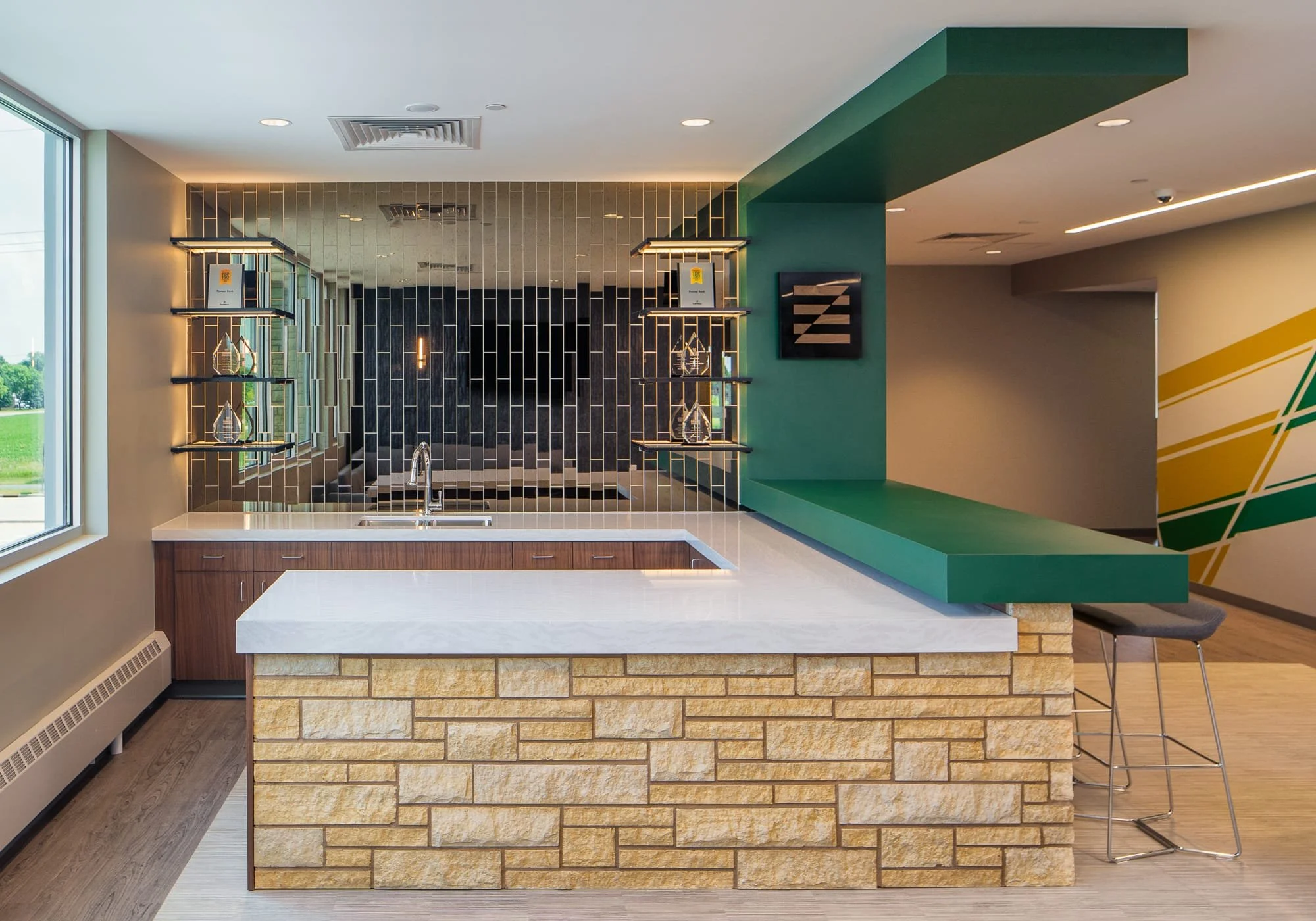 Contemporary commercial interior bar area with a stone-front counter, mirrored tile wall, green accent canopy, display shelves, and lounge seating.