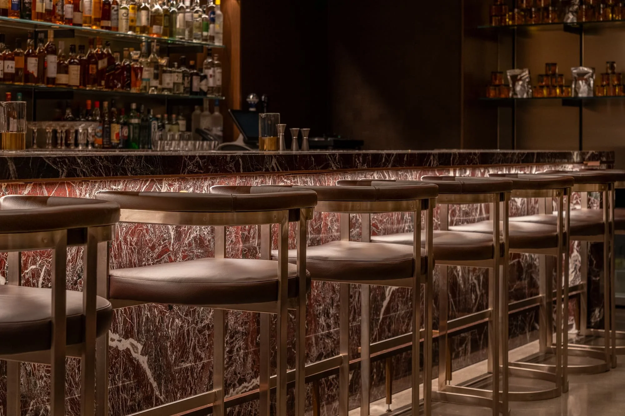 Row of leather bar stools along an illuminated marble counter with glass shelving and warm ambient lighting in a refined bar interior.