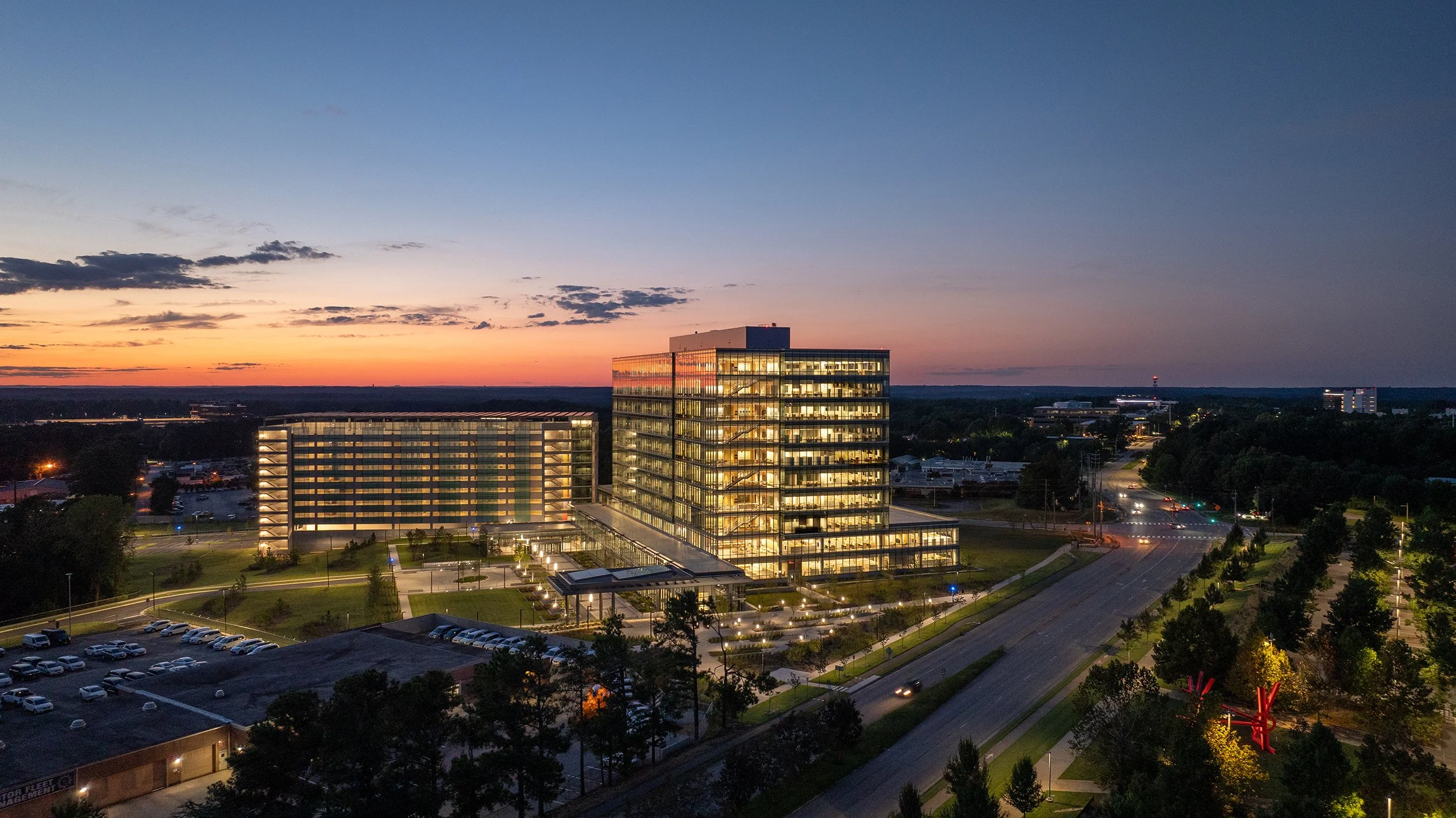Aerial dusk view of the illuminated DHHS campus with the office tower and parking deck silhouetted against a deep orange and blue sunset sky.