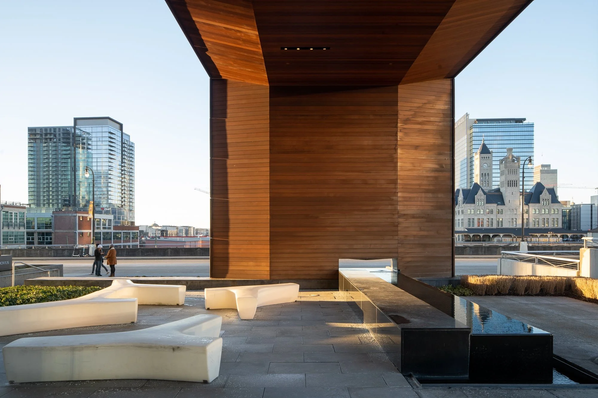 Gulch Crossing plaza level with the wood-clad pavilion cube, sculptural white benches, water feature, and the Nashville skyline including Union Station visible in the background