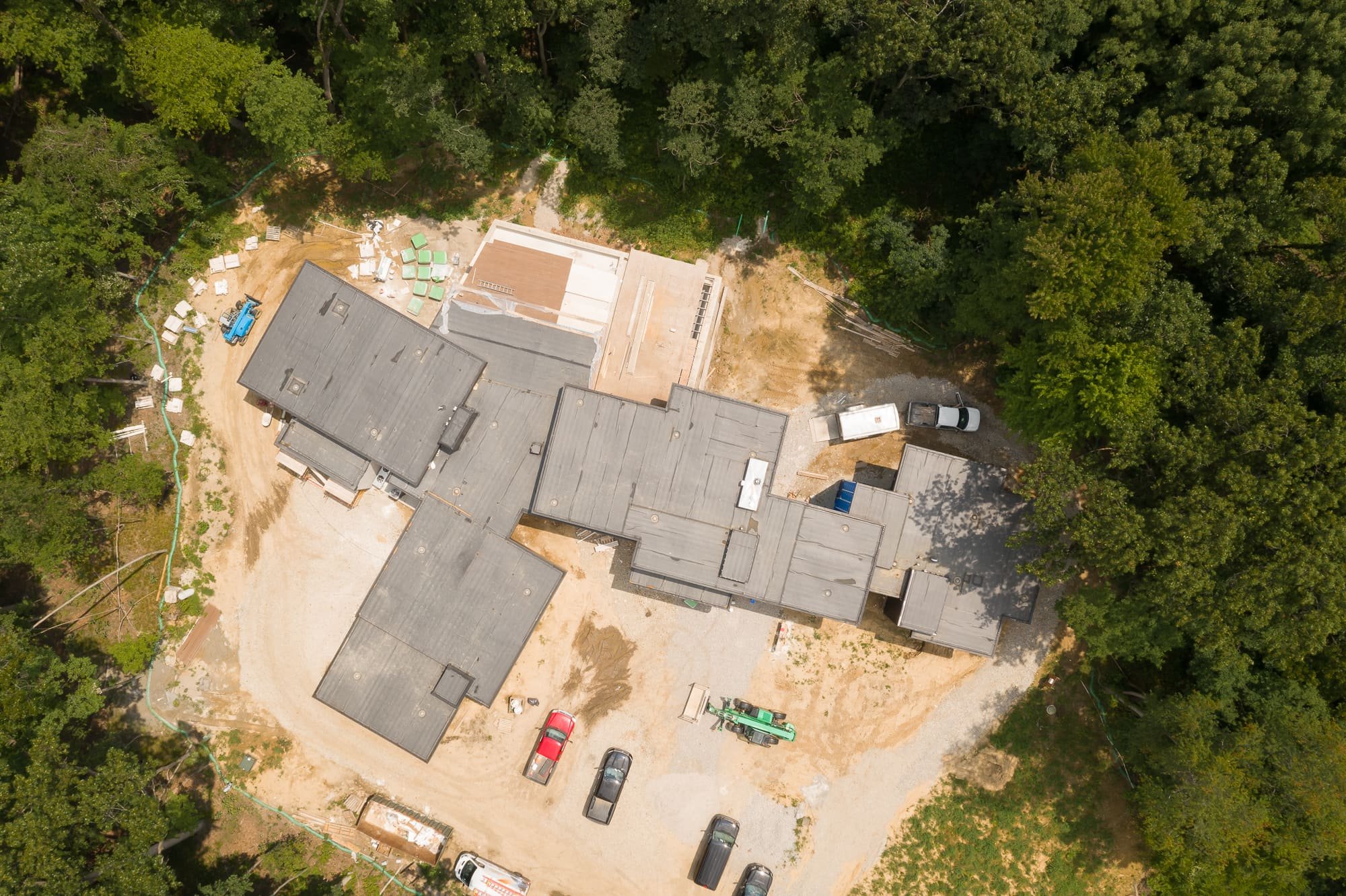 Aerial view of a residential construction site surrounded by dense trees, with vehicles, equipment, and building materials visible.