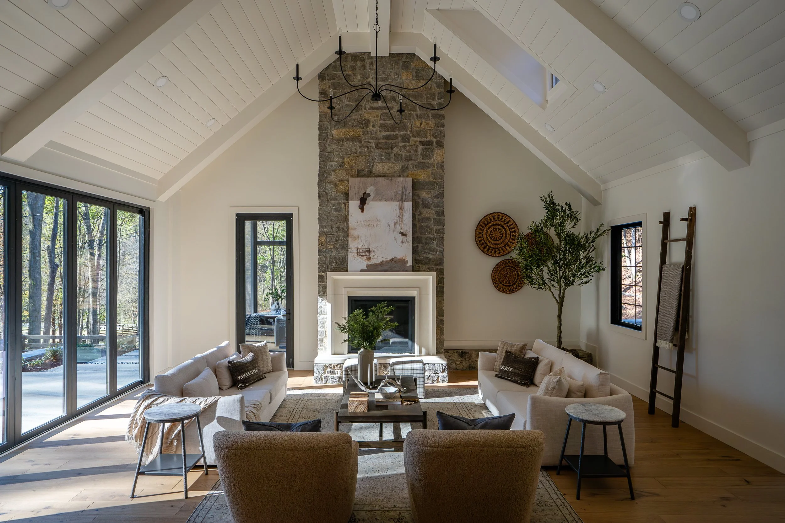 Vaulted living room with white beams, a tall stone fireplace, neutral seating, large glass doors, and natural light.